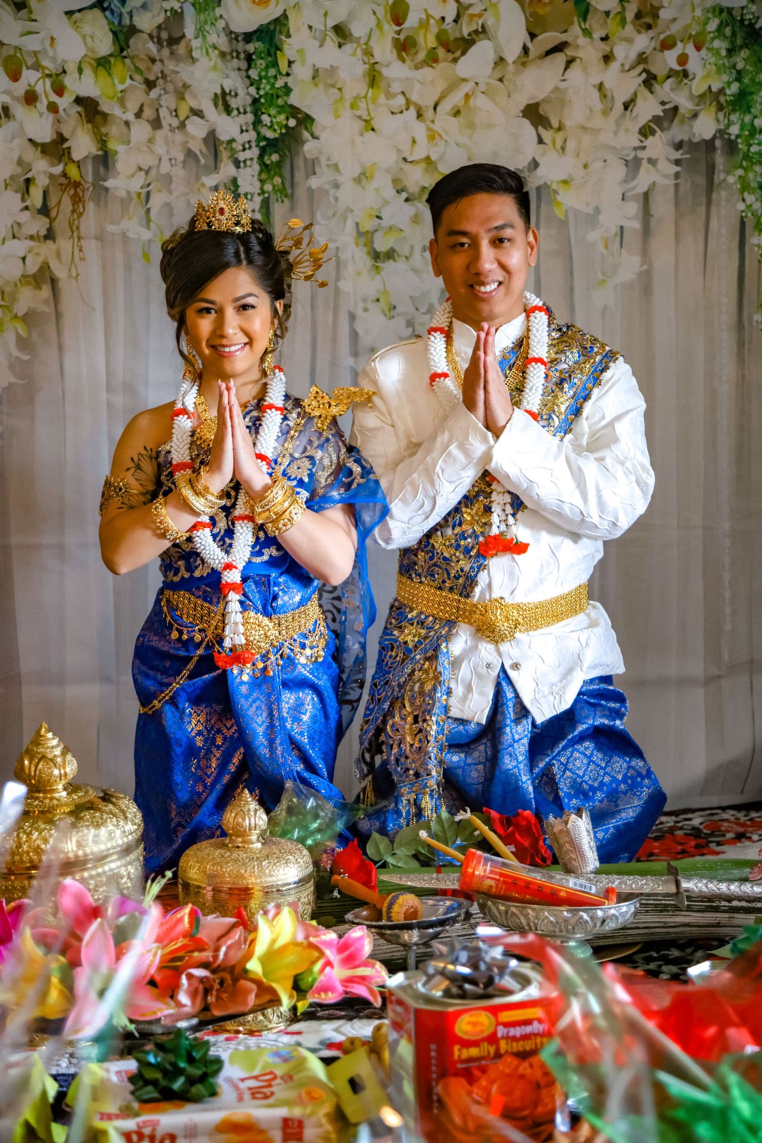 A couple in traditional Thai formal wear performing a ceremonial greeting with colorful decorations.