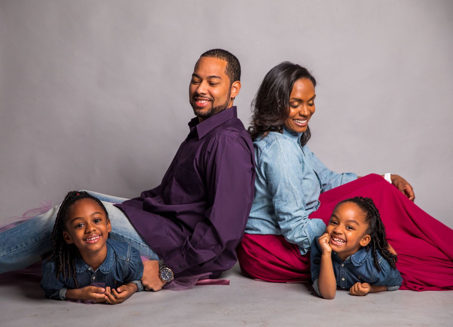 Happy Black family of four posing together on floor in coordinating outfits during professional studio photo session.