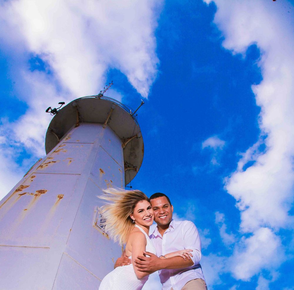 A couple shares an intimate moment in front of a white lighthouse under vibrant blue skies.