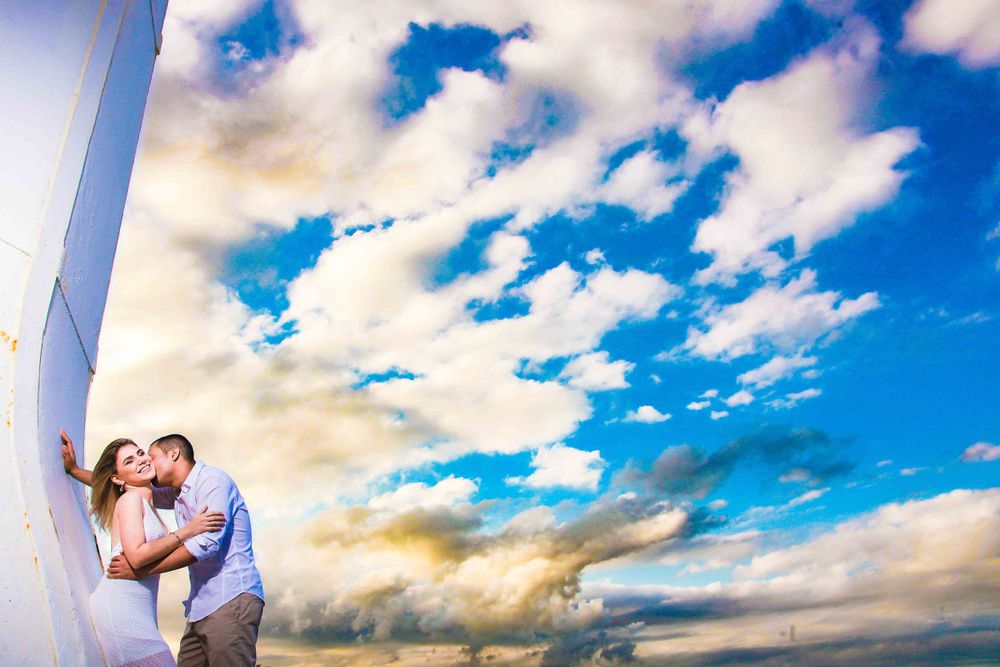 A romantic couple embraces against a dramatic sky with white clouds and a lighthouse in the background.
