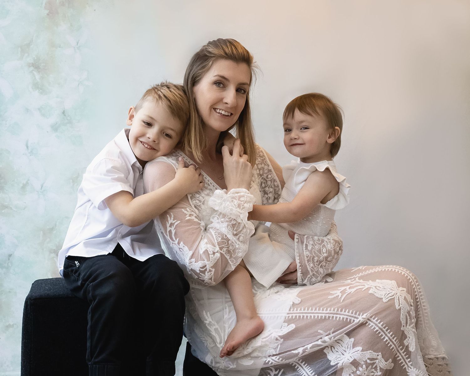 A family portrait with mum in a white lace dress embracing two young children against a soft grey backdrop.