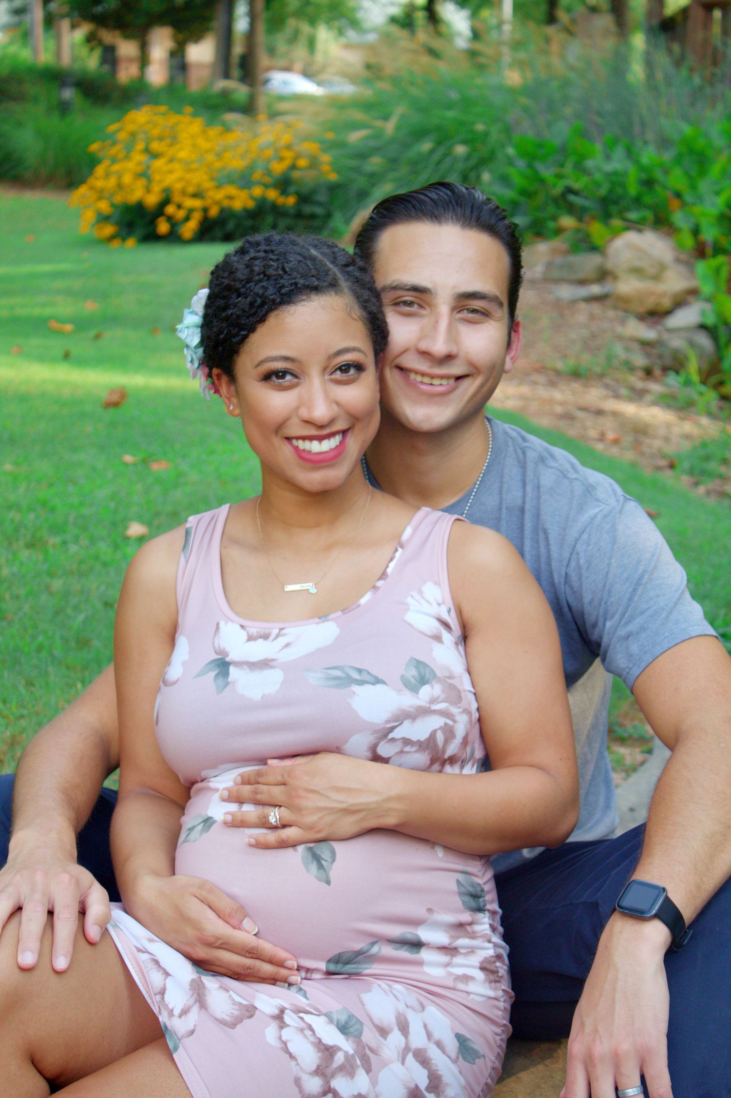Maternity photo of a couple in casual clothes sitting outdoors in a garden setting.