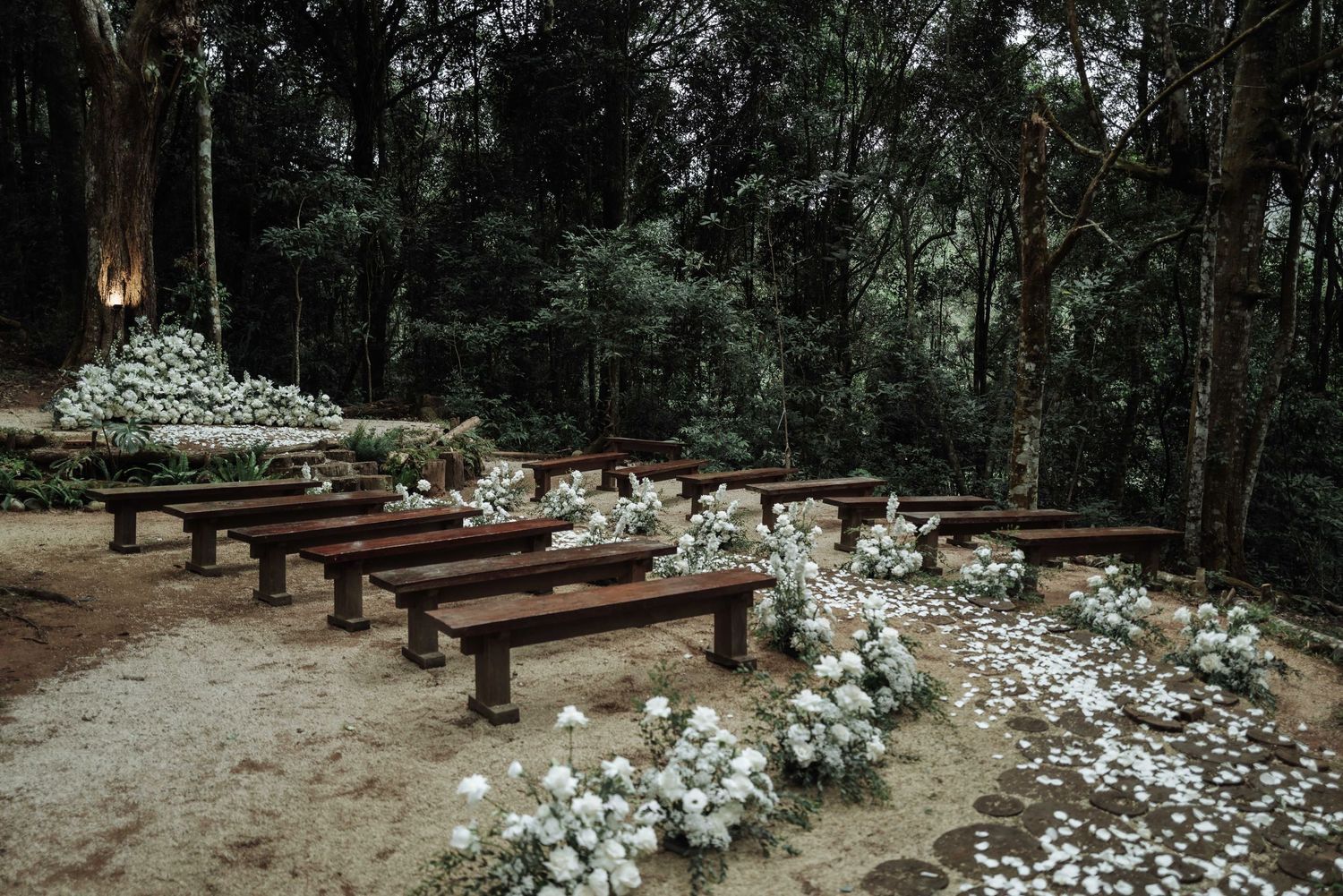 Rustic outdoor wedding ceremony setup with wooden benches lined with white floral arrangements in a forest.