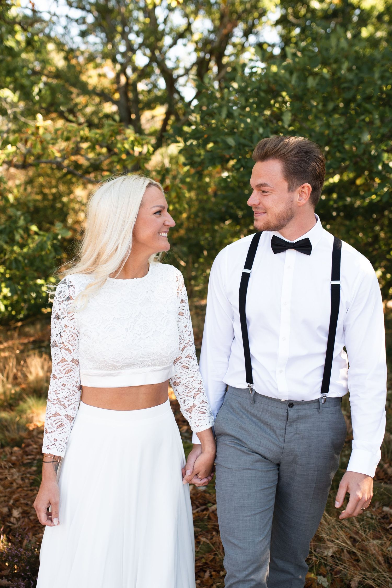 Stylish couple in wedding attire with suspenders and crop top lace dress in sunlit garden.