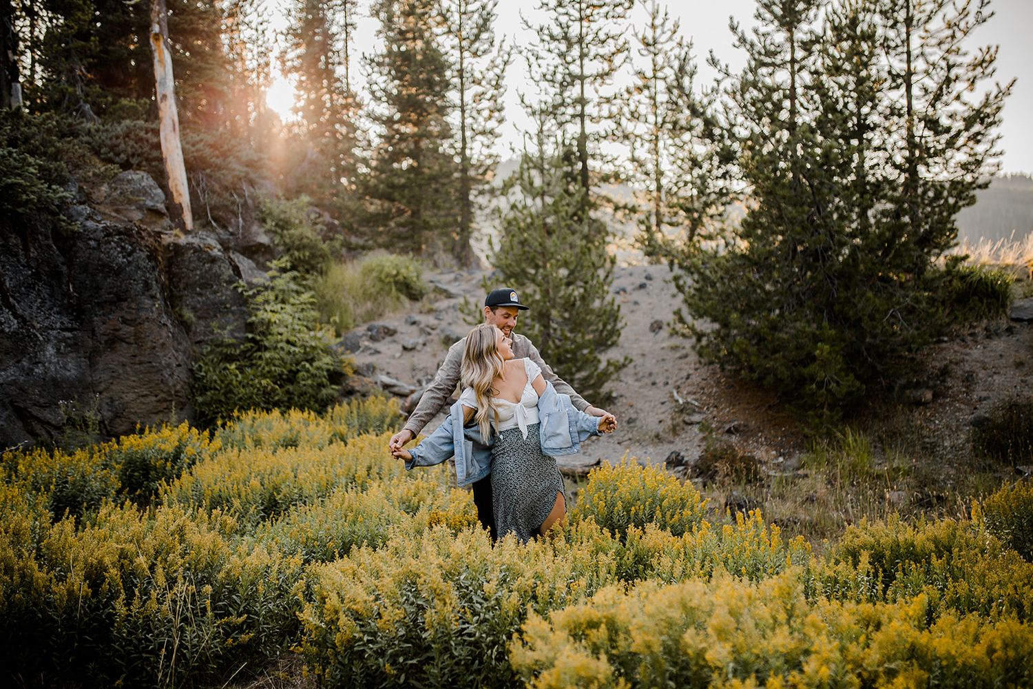 Figure standing among yellow wildflowers on a mountainside with sunlit pine trees in the background.