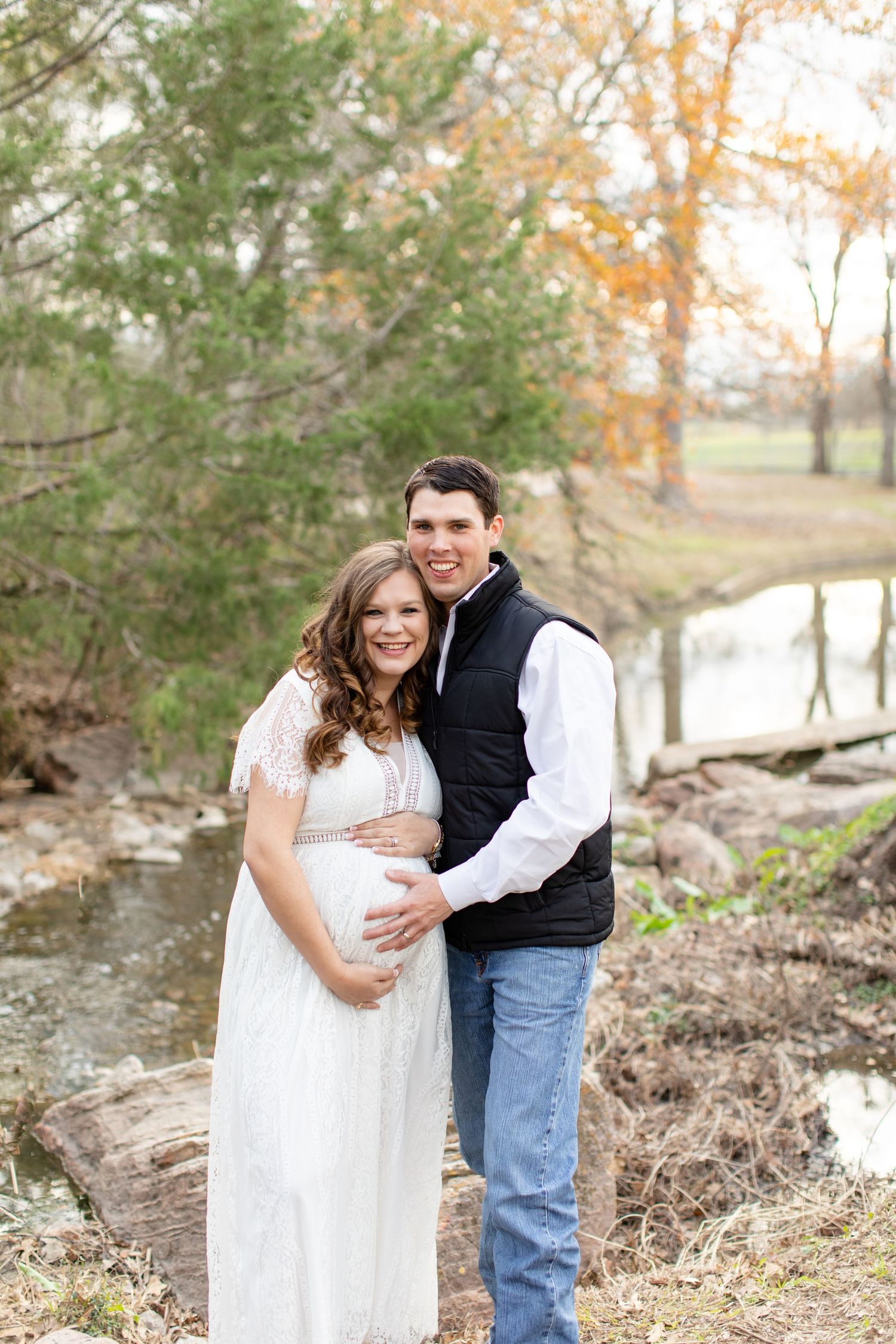 Maternity photo of couple in white dress and vest standing by creek during autumn.