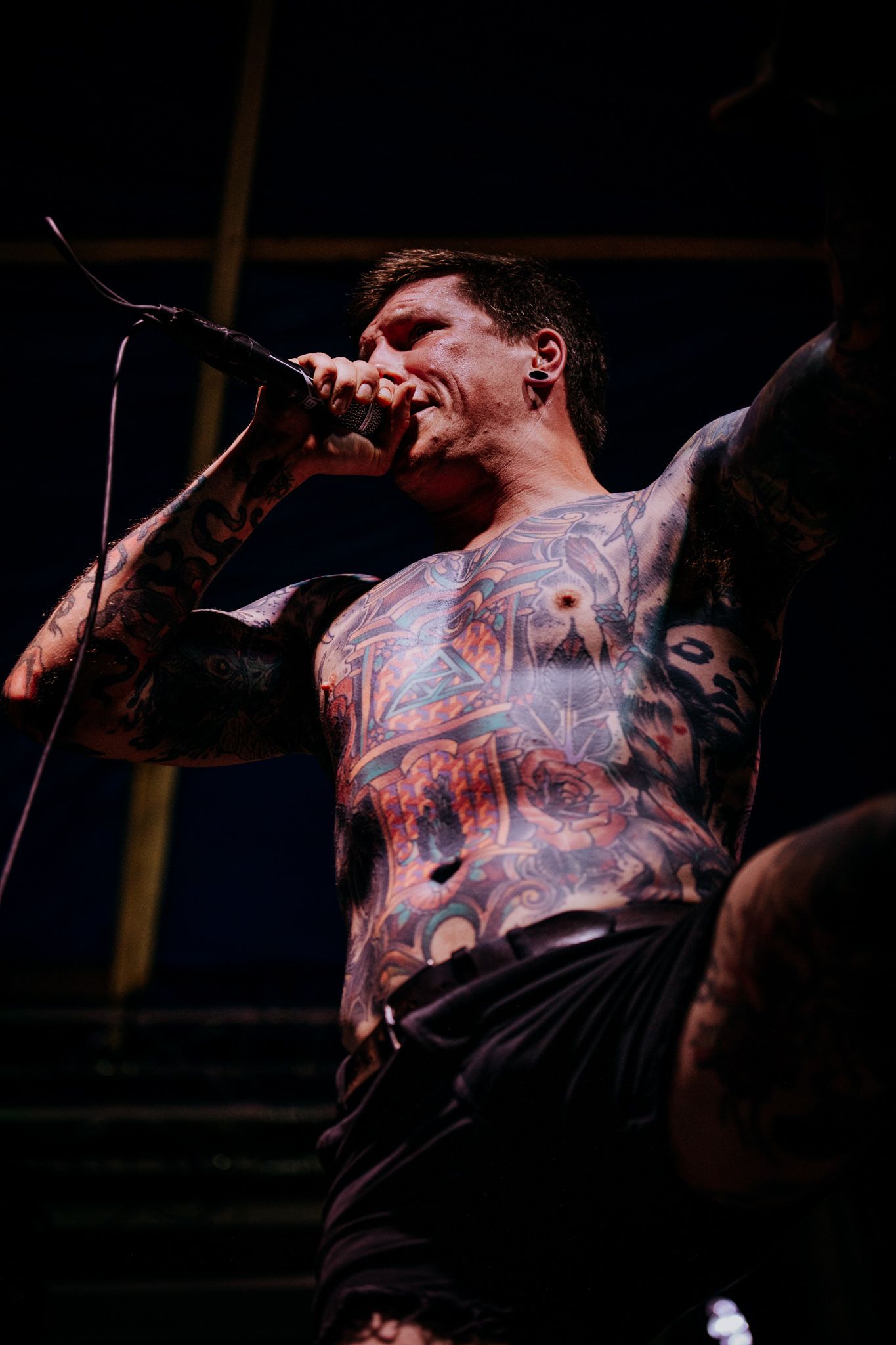Tattooed performer singing passionately on stage during dark concert performance.