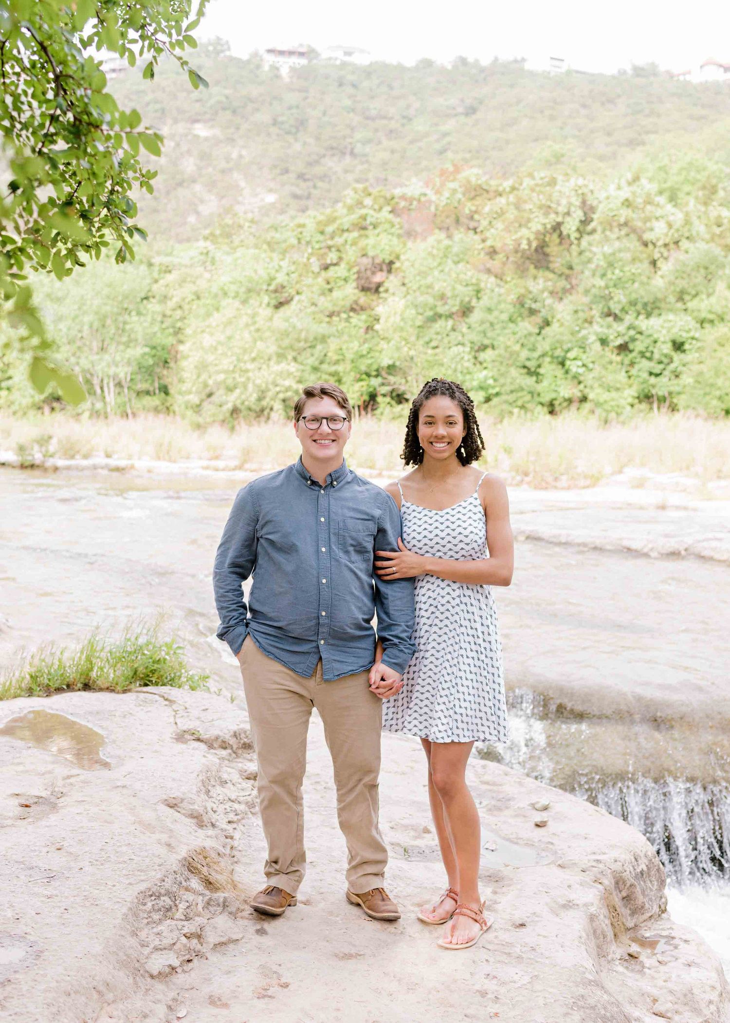 Couple standing together by a scenic riverside with lush greenery in the background.
