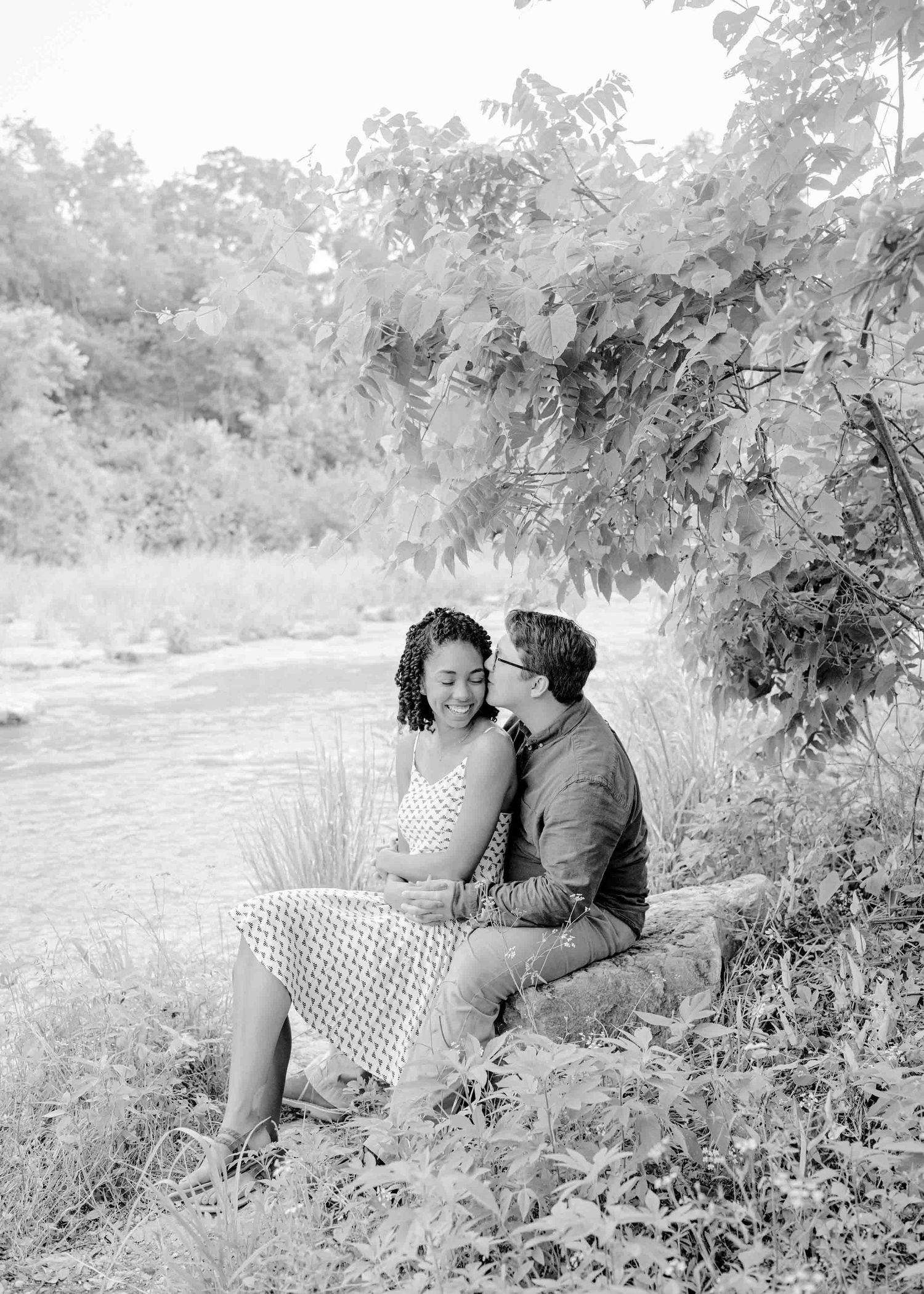 Black and white photo of a couple sitting together by a serene riverside.
