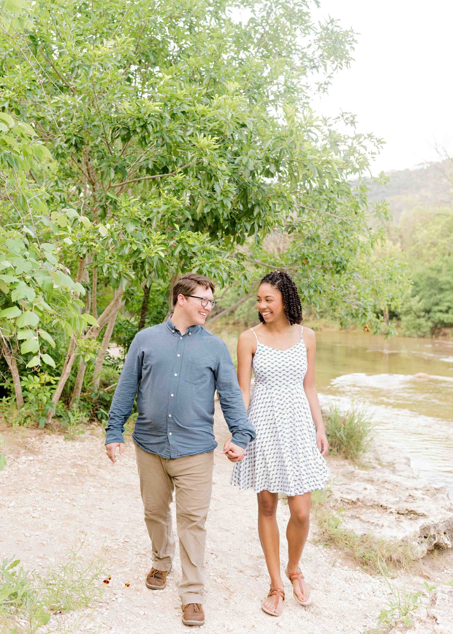 Couple walking hand in hand along a riverside path surrounded by lush greenery.
