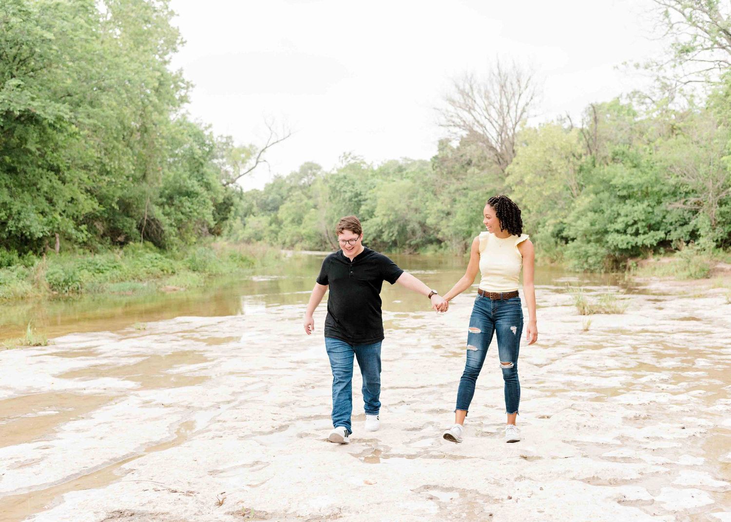 A couple holds hands while walking together on rocky terrain near a river on a sunny day.