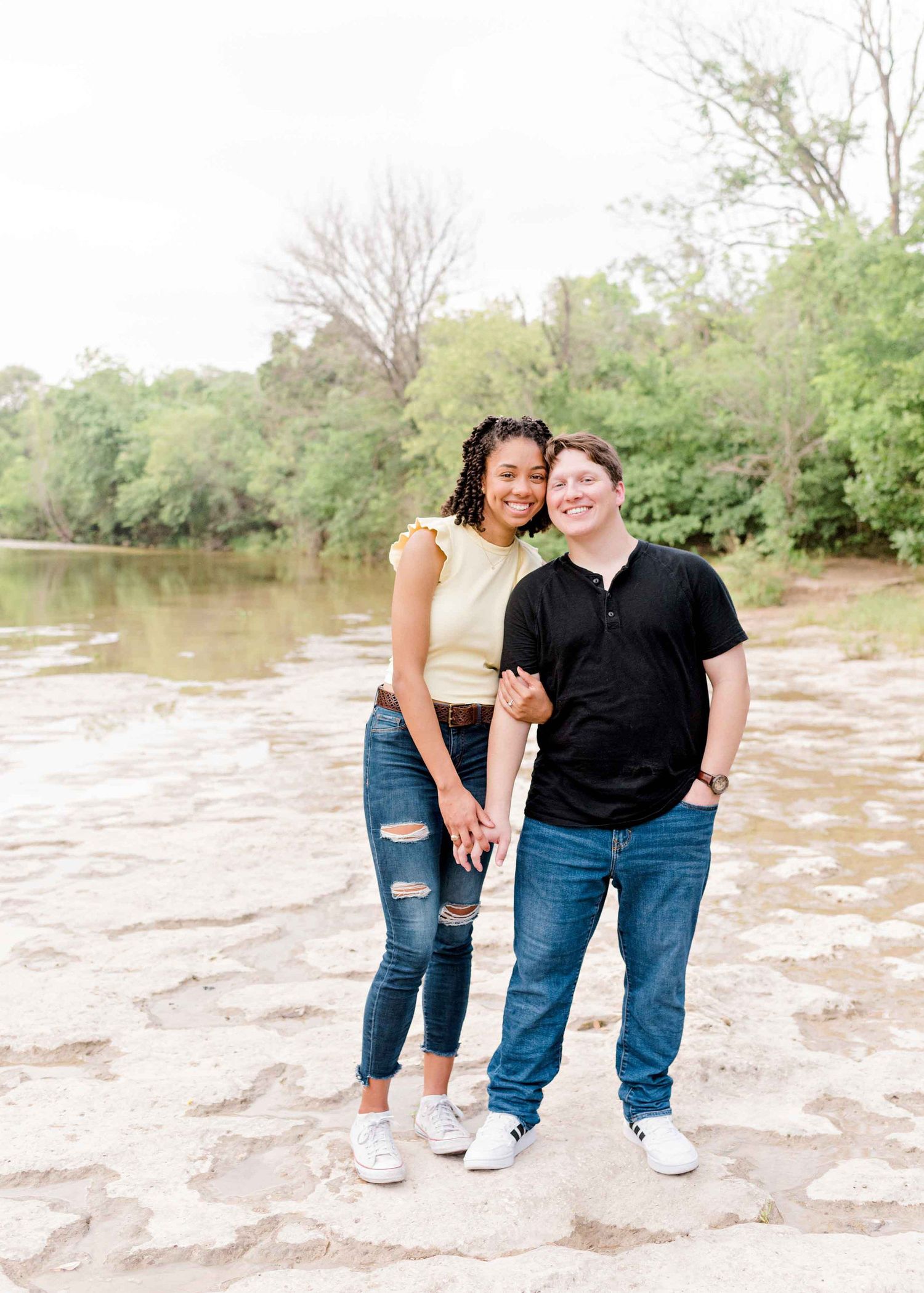 A couple stands together on rocky terrain near a river surrounded by green foliage.