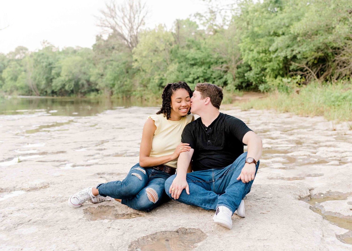 A couple sits together laughing on rocky terrain near a river during their engagement session.