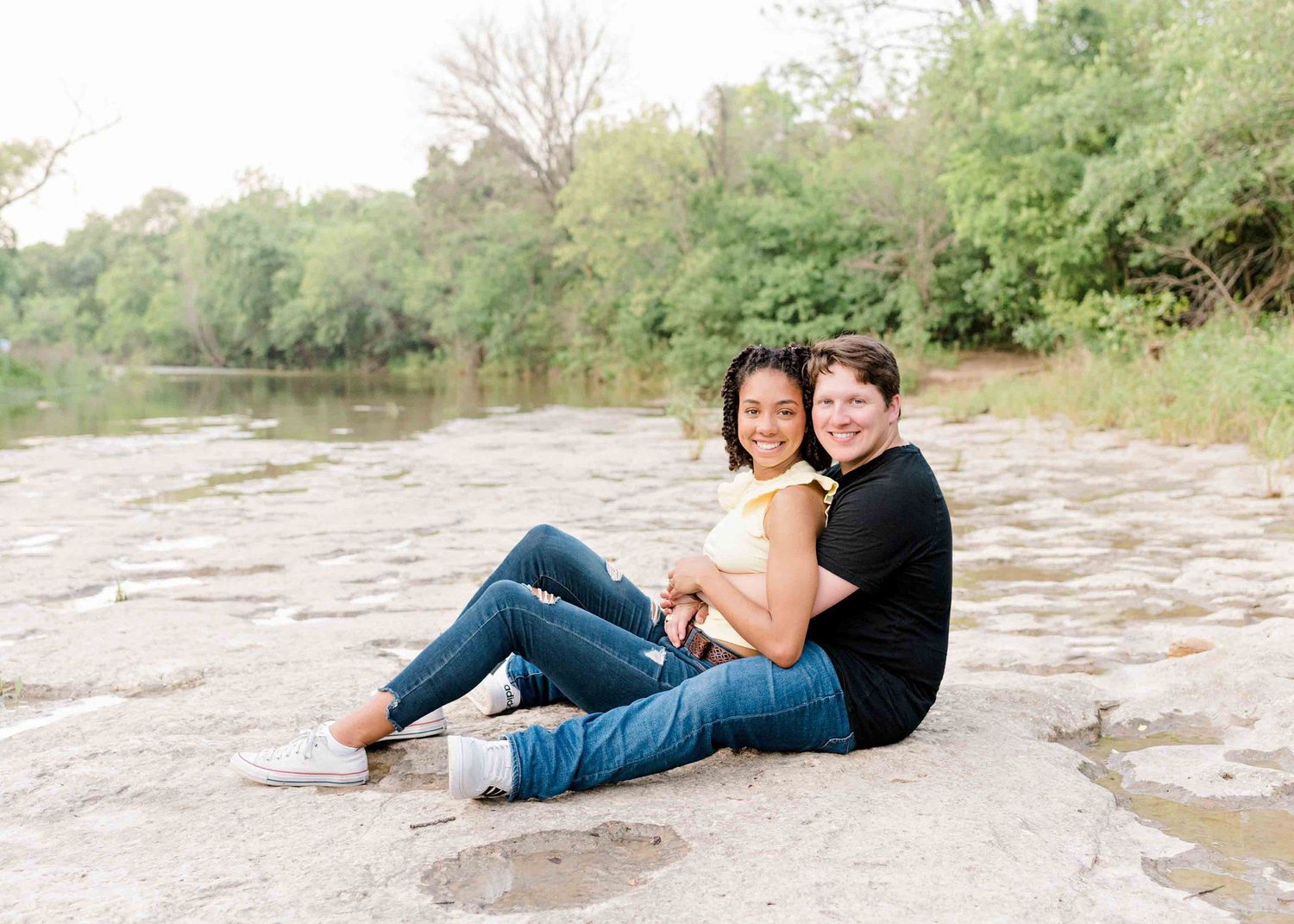 A couple sits together on rocky terrain near water and trees on a sunny day.