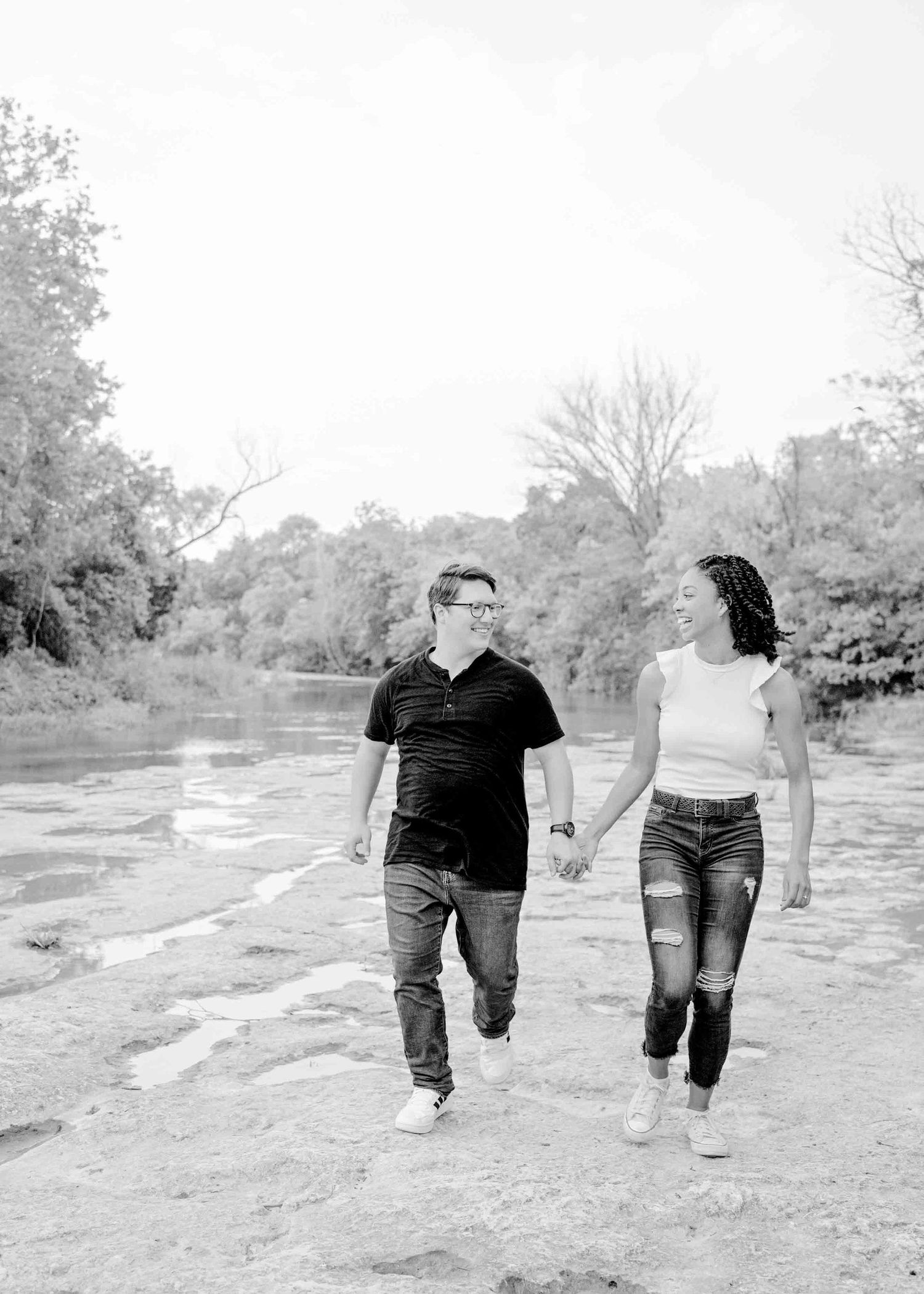 A couple walks hand in hand along a rocky riverbank on a sunny day.