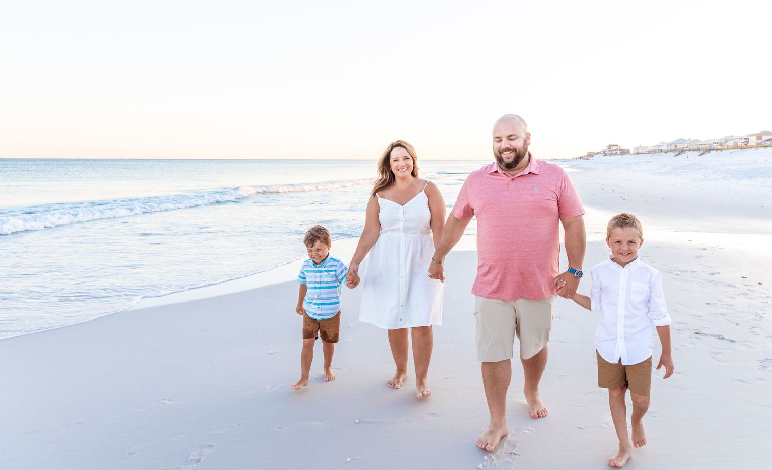 Family of four walking together on white sandy beach during sunset.