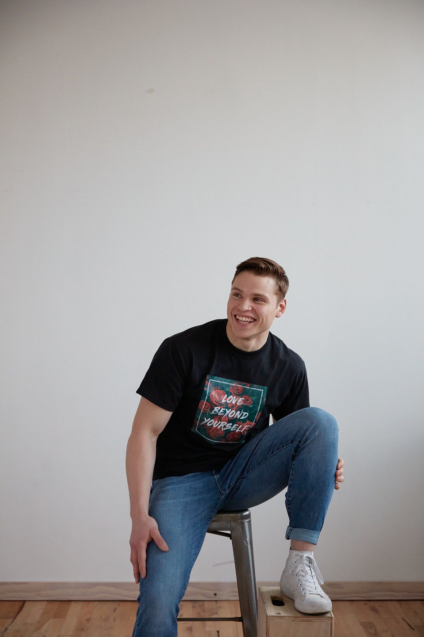 A casual portrait against a white wall featuring a person seated on a stool wearing jeans and a graphic t-shirt.