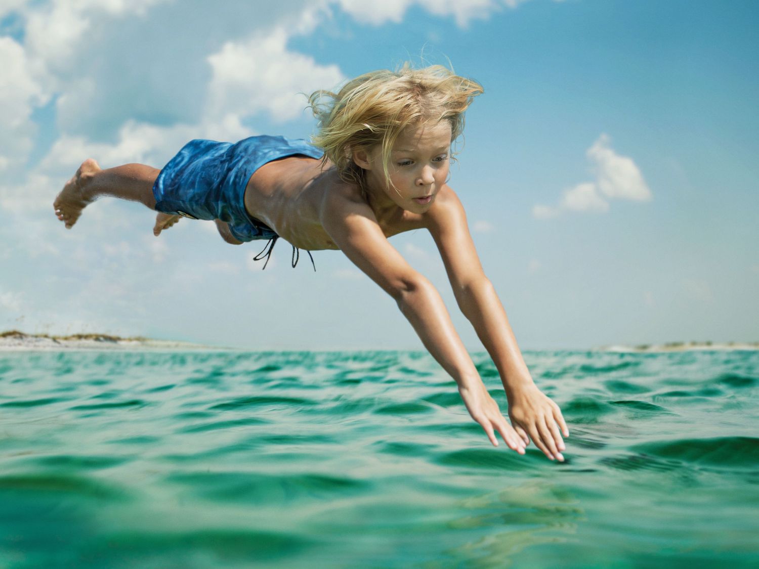 Blonde child diving into turquoise ocean water with blue shorts on a sunny day.