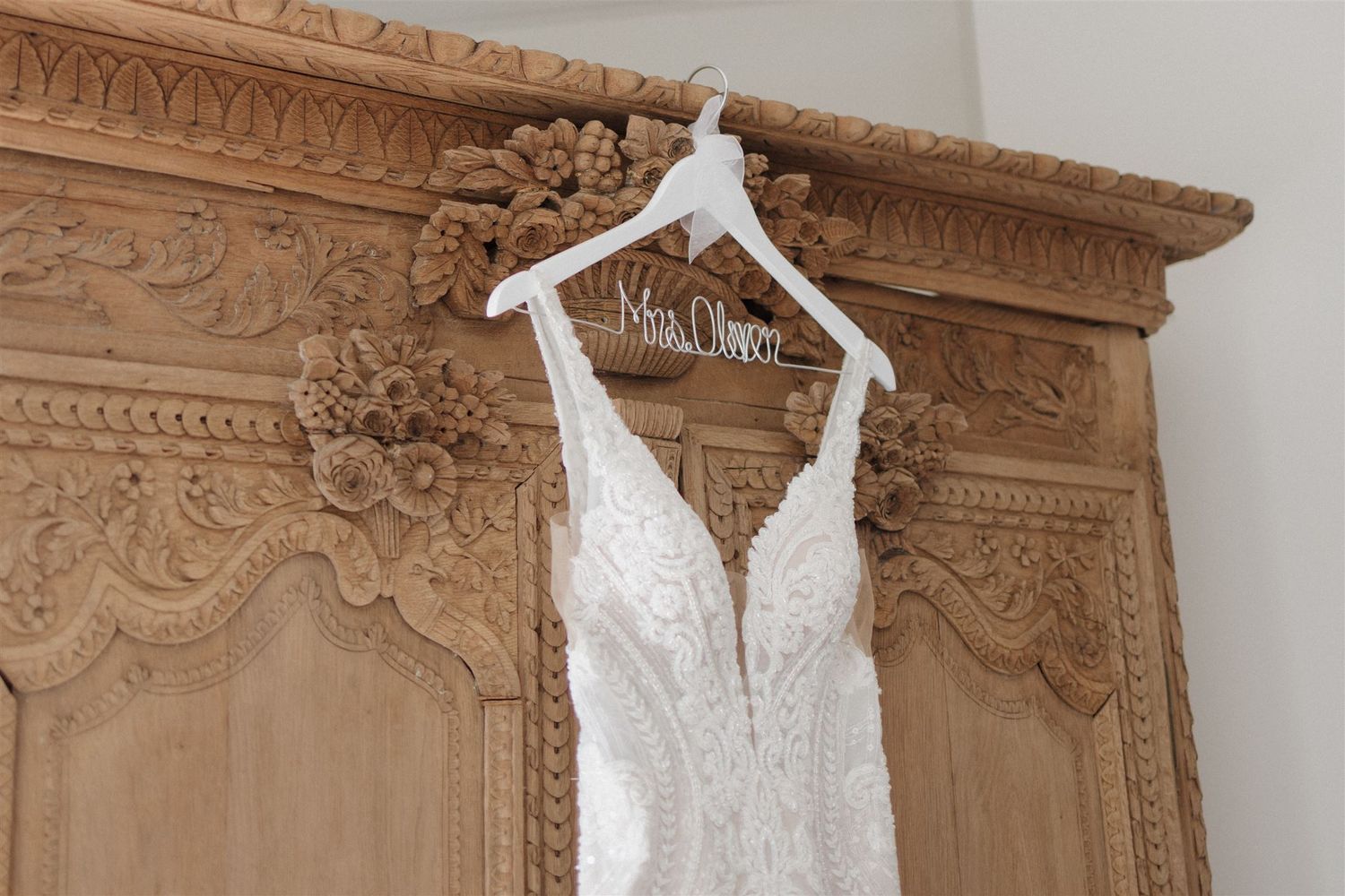 Delicate lace wedding dress hangs on white hanger against ornate wooden wardrobe.