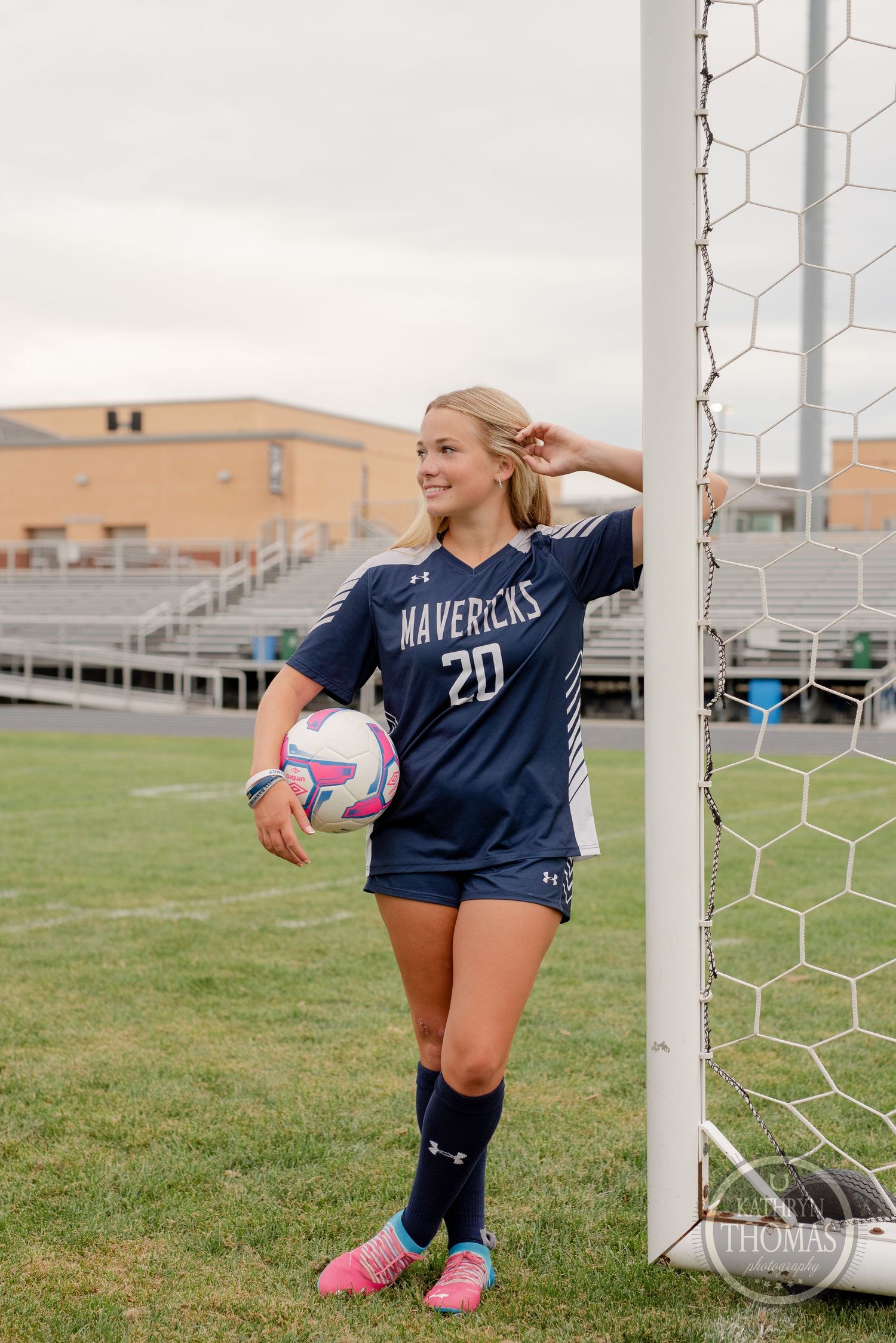 Soccer player in navy Mavericks jersey #20 poses with ball by goal post on field.