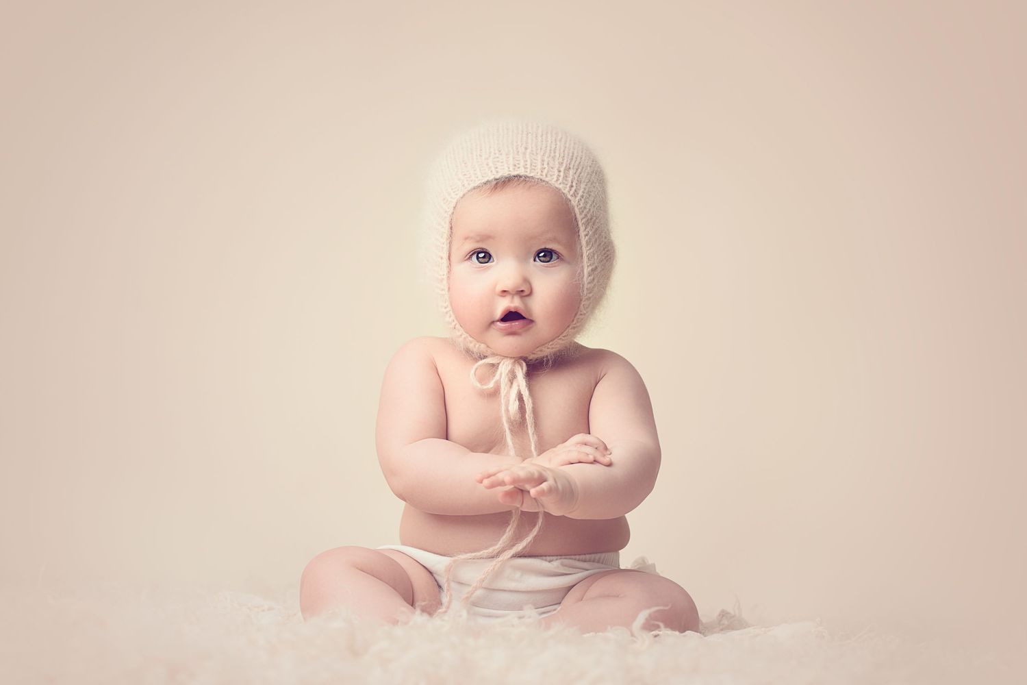 Baby in white knit bonnet sitting on soft cream backdrop during professional portrait session.