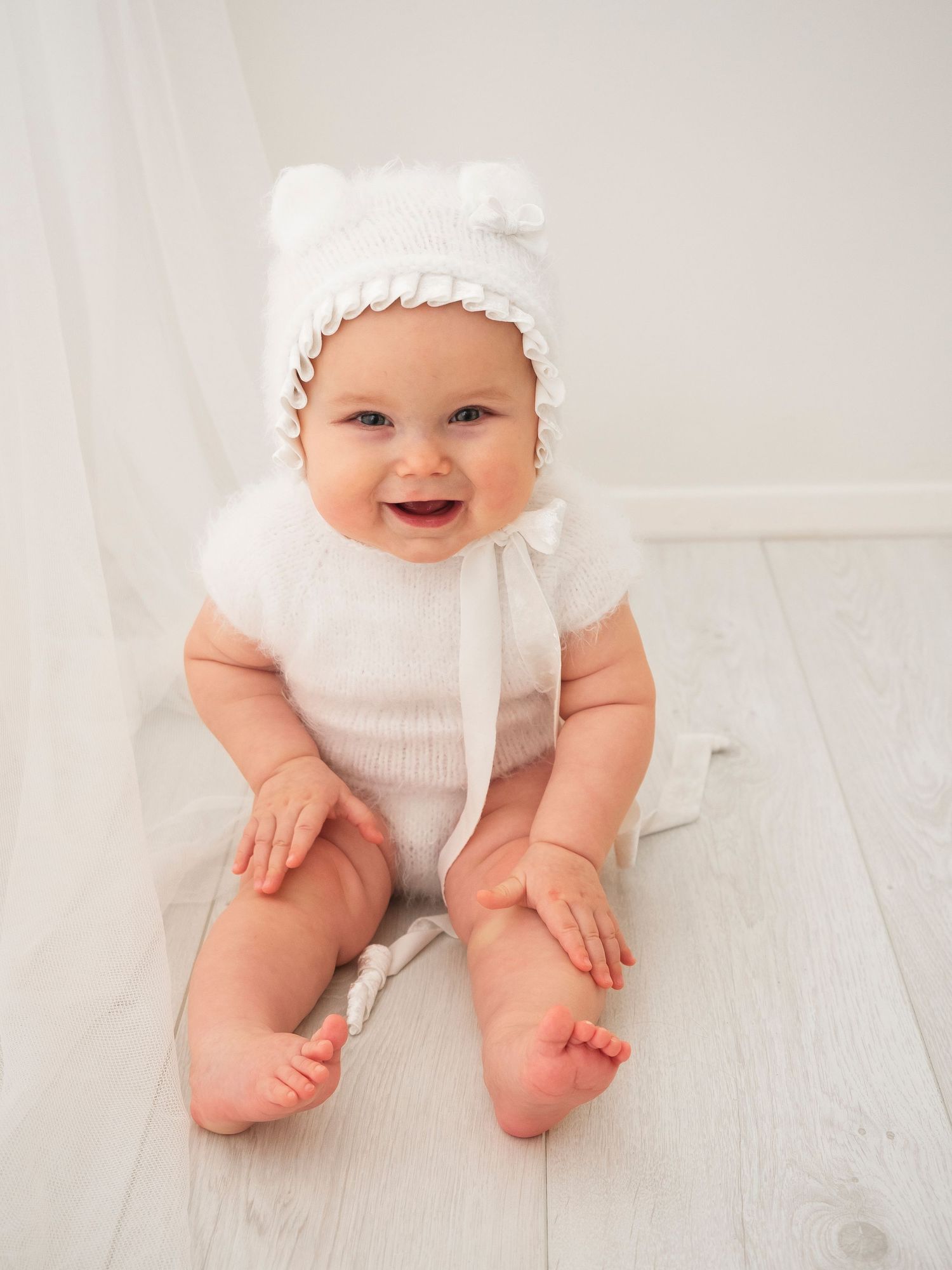 Baby girl in white vest with matching bonnet laughs for her photoshoot.