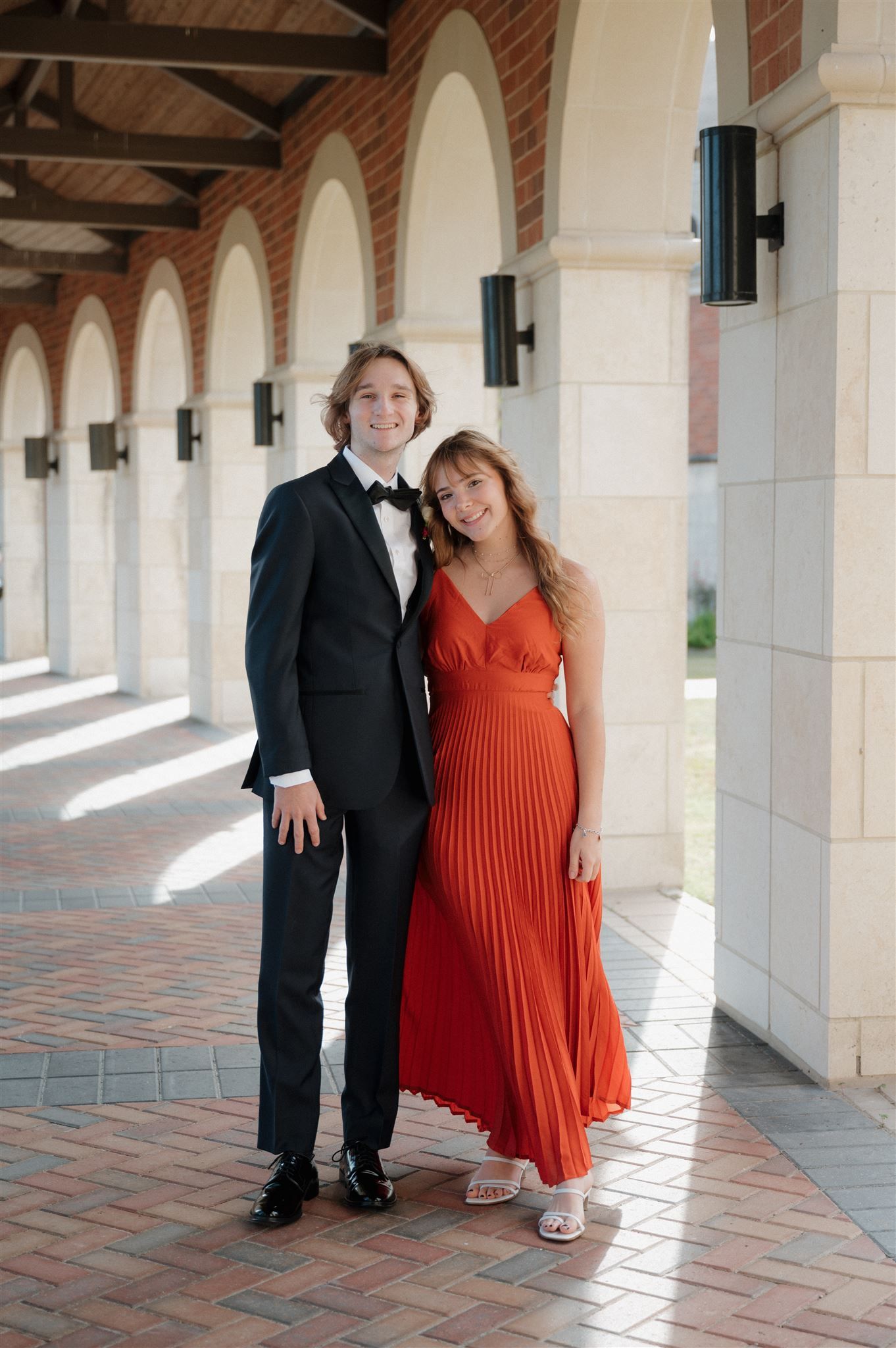 Formal couple poses in black tie and red dress under an ornate columned walkway.