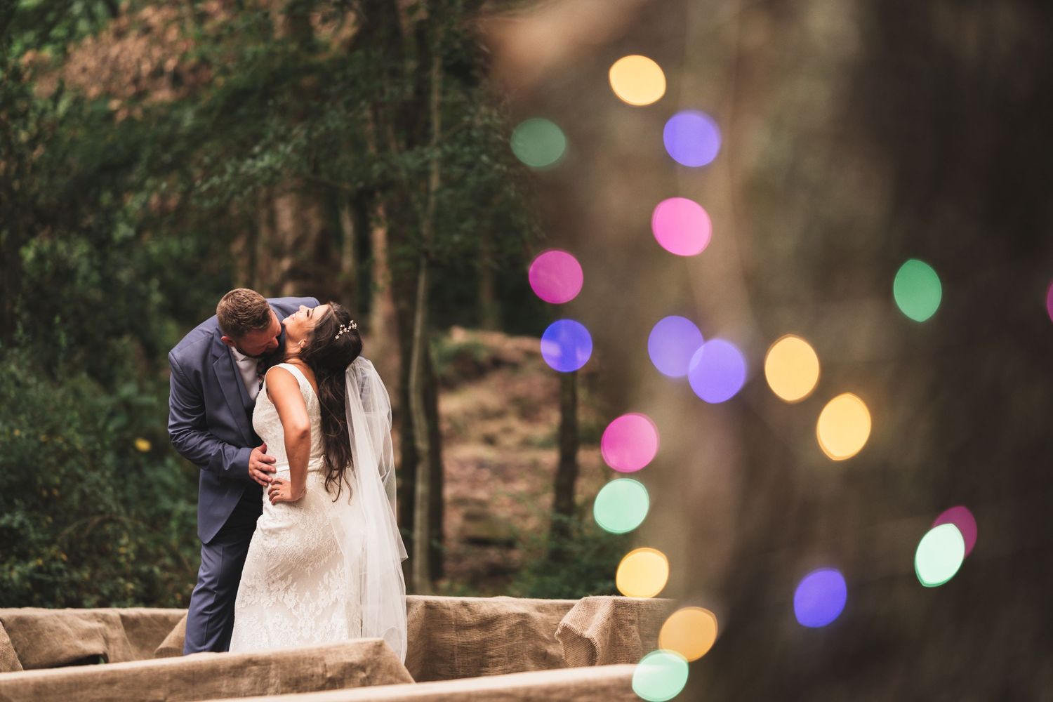 Bride and groom sharing a kiss surrounded by colorful bokeh lights in a lush outdoor setting.