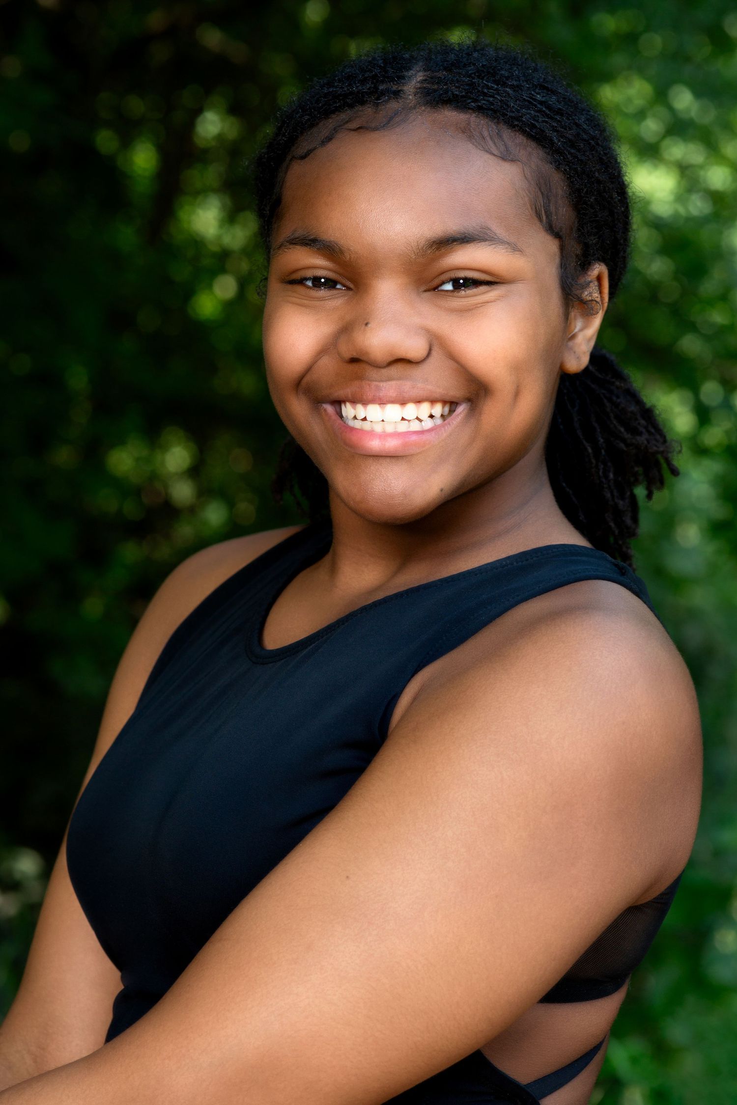 An outdoor portrait of a smiling person in a black tank top against lush green foliage.
