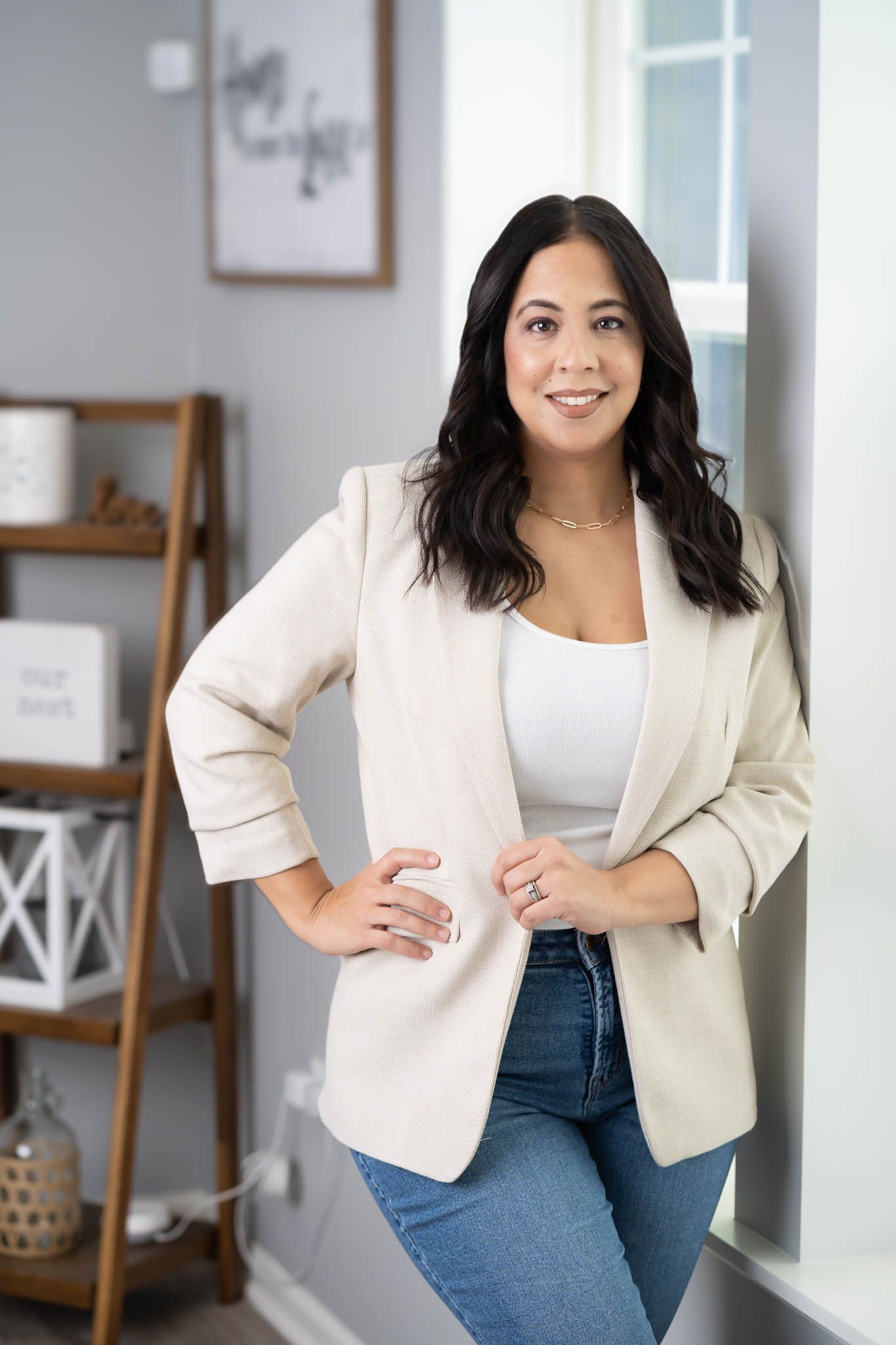 Professional pose in cream blazer and jeans against modern home interior.