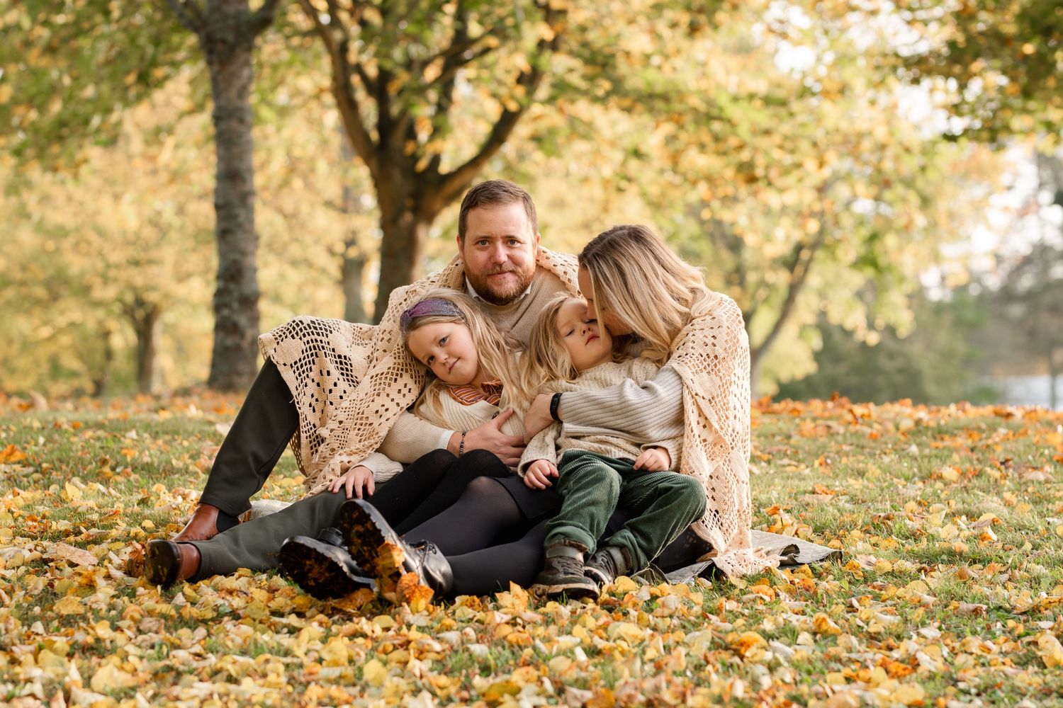 Family of four cuddling together on autumn leaves in a park setting.