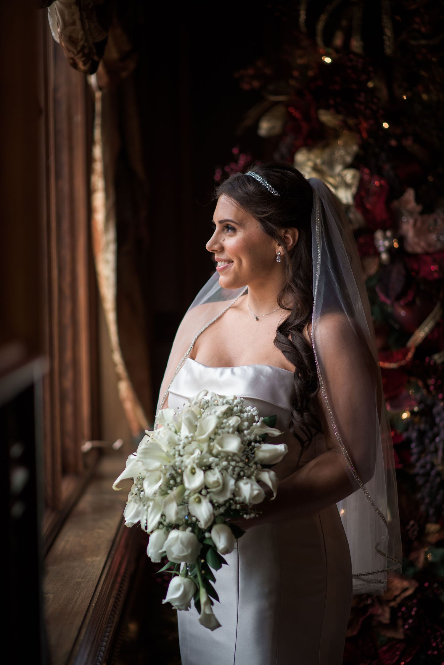 Beautiful bride in strapless gown holds white calla lily bouquet beside holiday decorations.