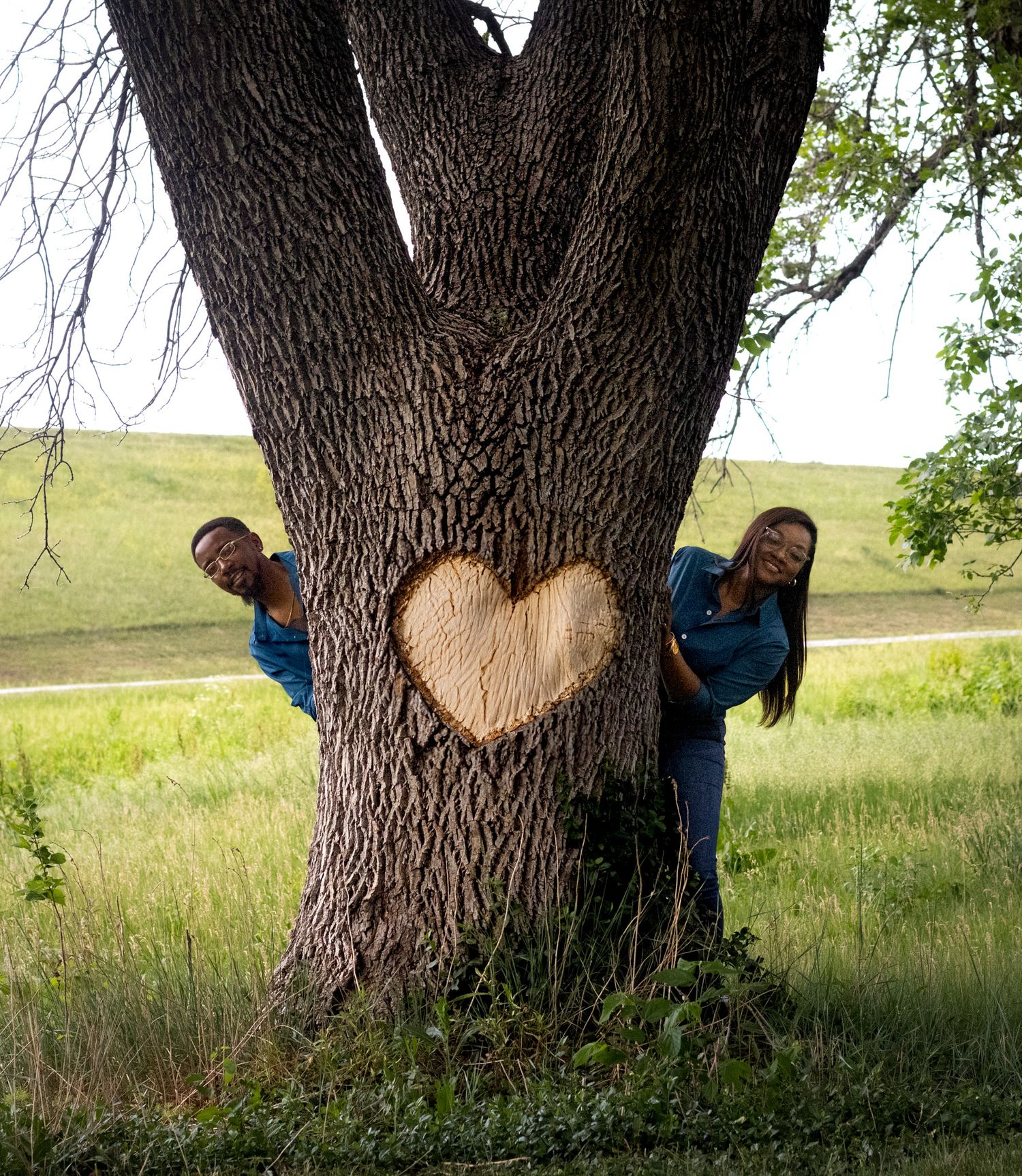 Two people peek playfully from behind a large tree trunk with a heart carved into its bark in a grassy field.
