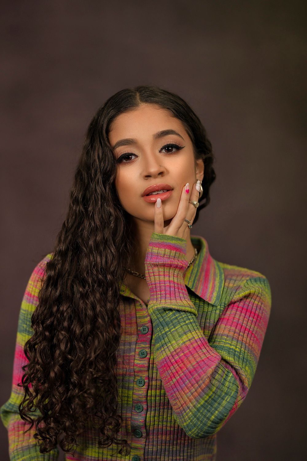 Fashion model wearing a vibrant pink and green knit sweater poses with flowing curly hair against dark background.