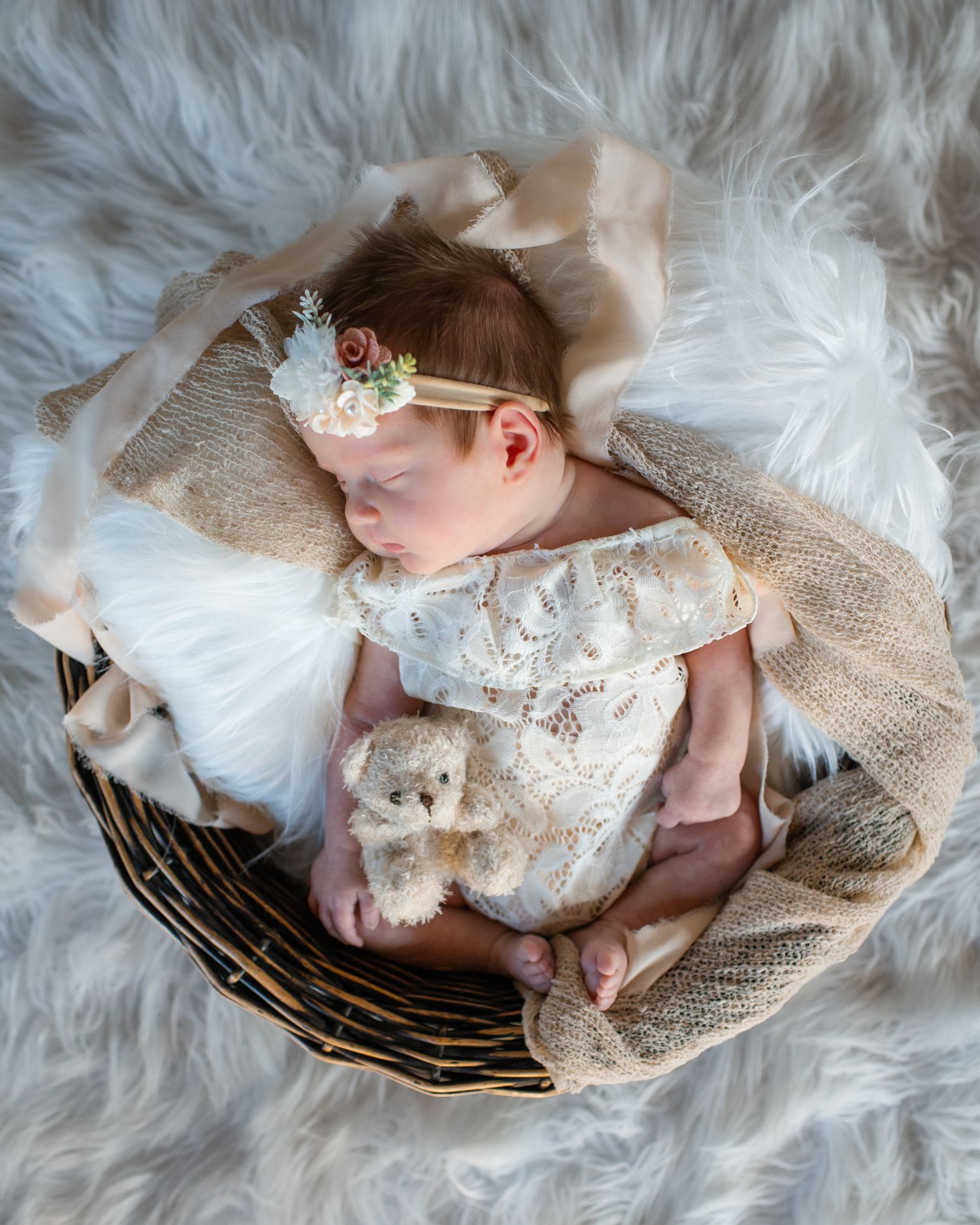 Sleeping newborn in lace dress and headband rests in basket on fluffy white backdrop.