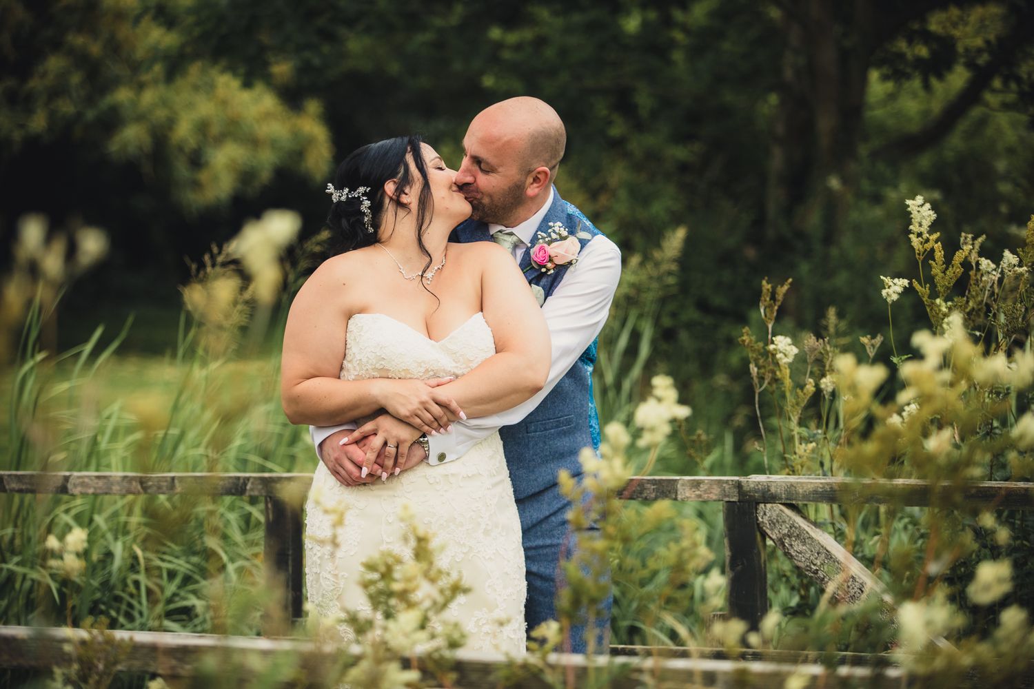 bride and groom stood on bridge kissing arms around each other