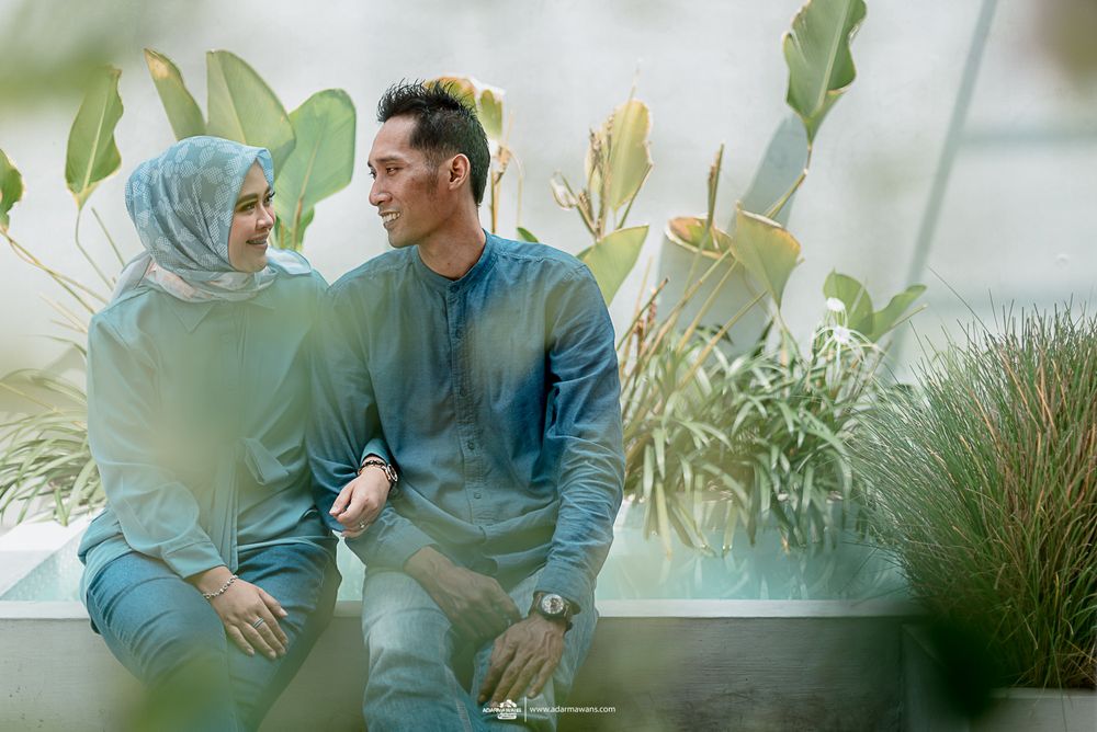 Couple in coordinating blue outfits pose amid green foliage and soft lighting.