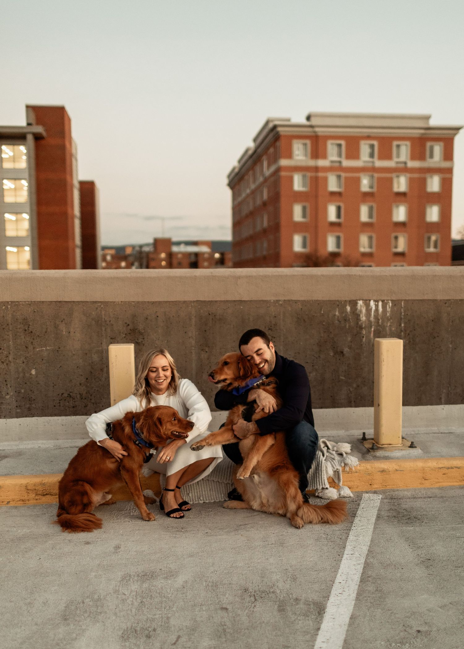 Couple sitting with their two dogs on rooftop parking deck with brick buildings in background.