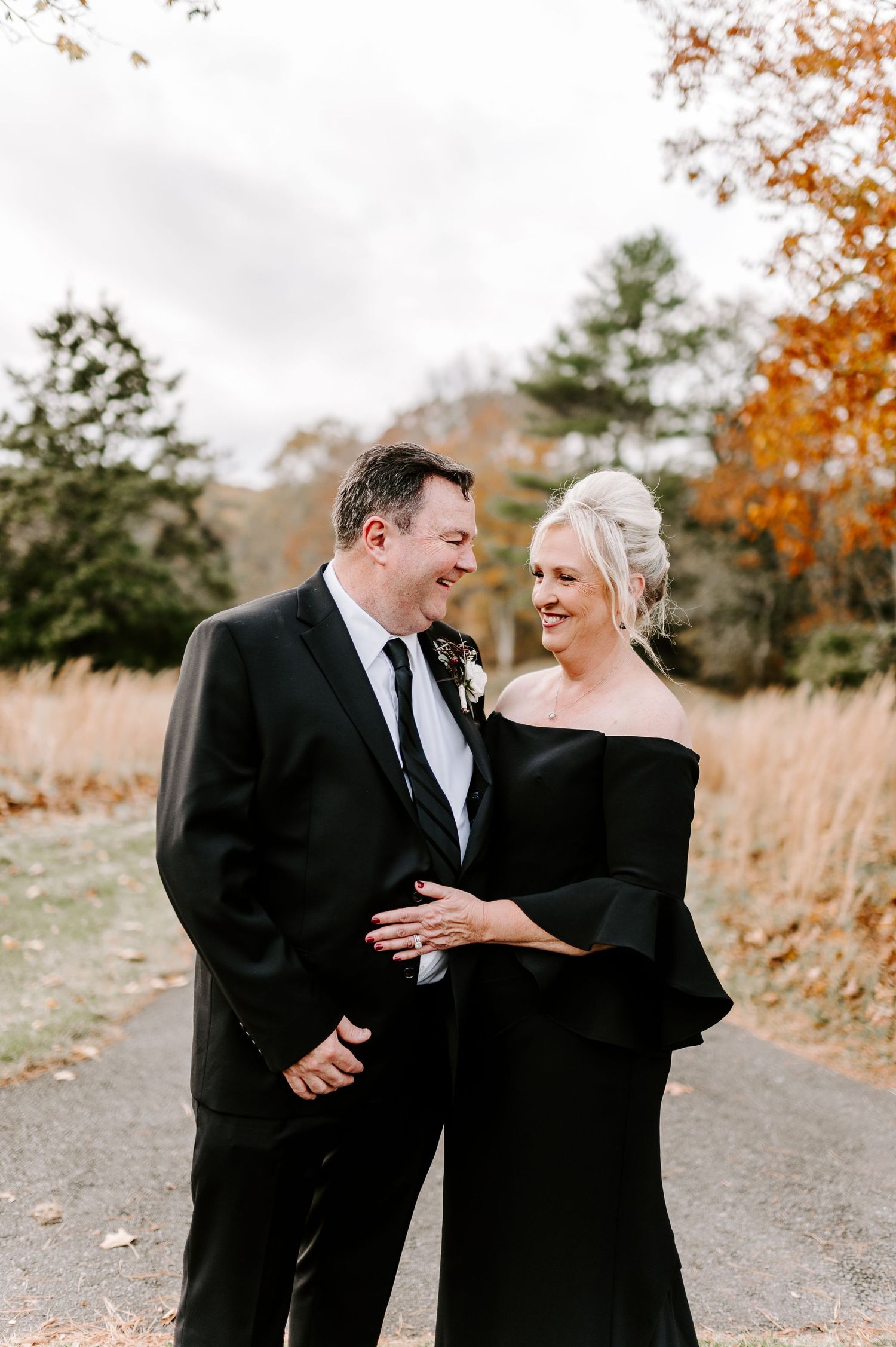 Couple in formal black attire shares candid laughs during outdoor autumn photoshoot.