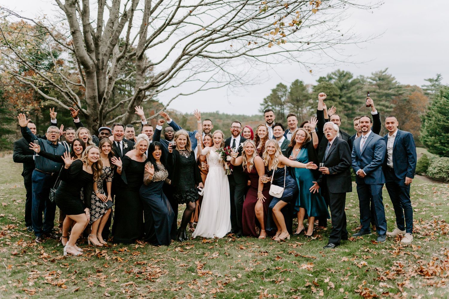 Large wedding party poses outdoors in autumn with colorful fall foliage in the background.
