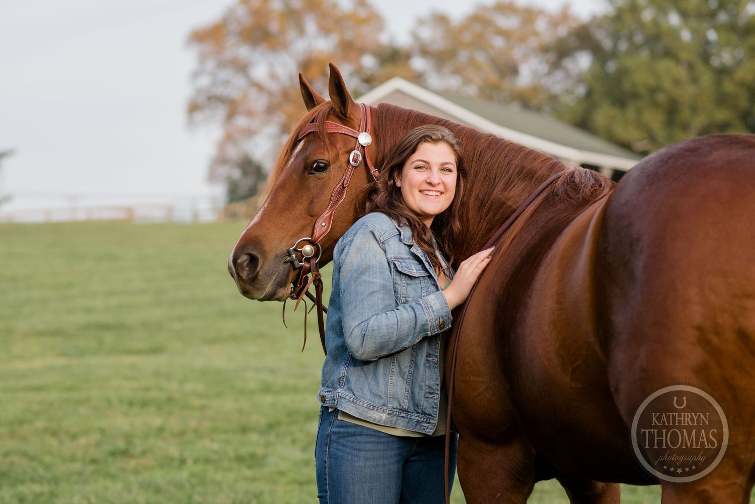 A denim-clad figure stands close to a chestnut horse in an autumn field.