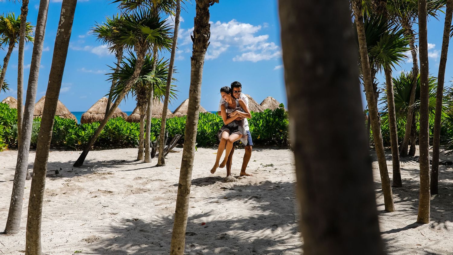A couple embraces under palm trees on a tropical beach with white sand and blue skies.