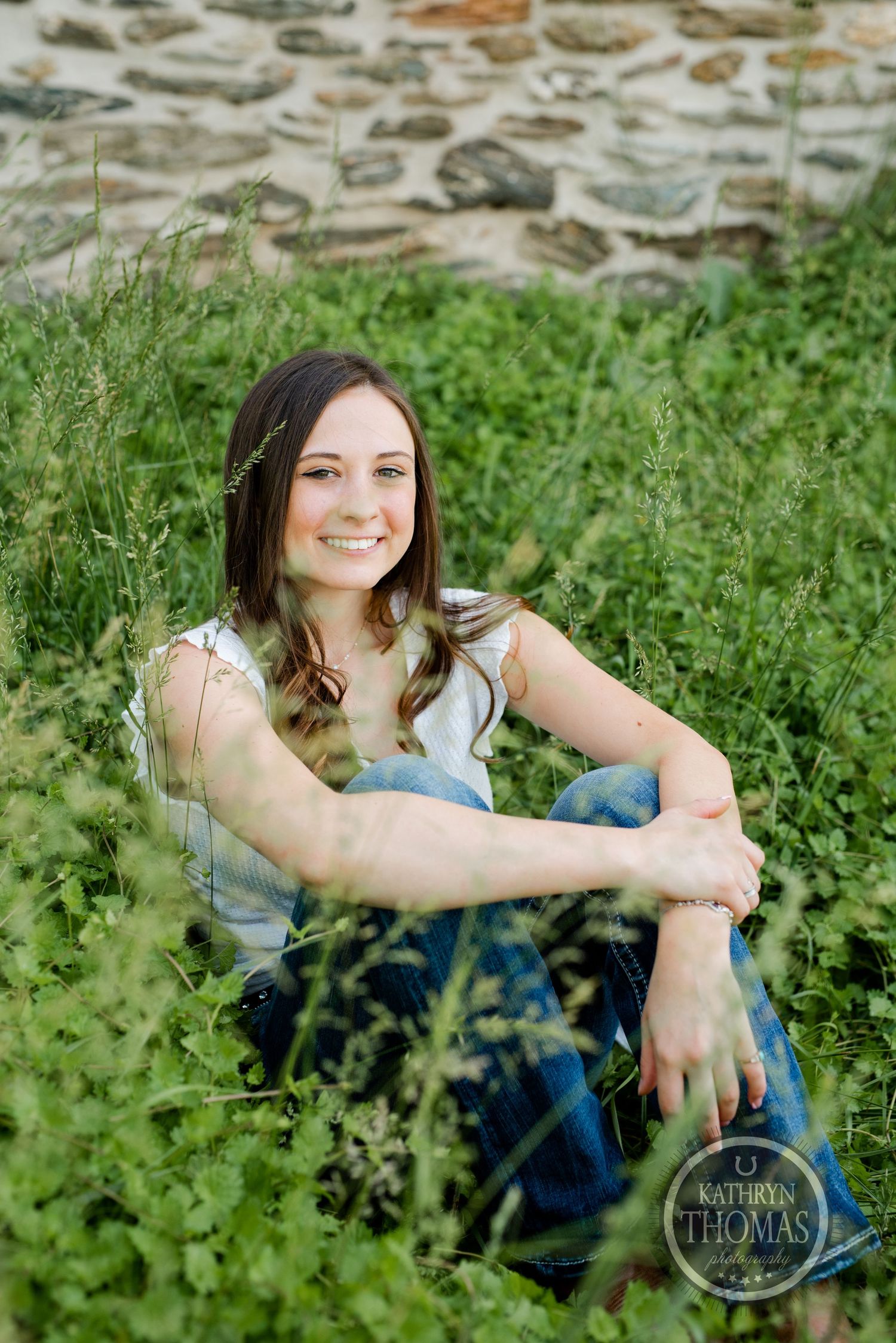 A portrait session in green foliage against a stone wall backdrop wearing casual denim and white top.