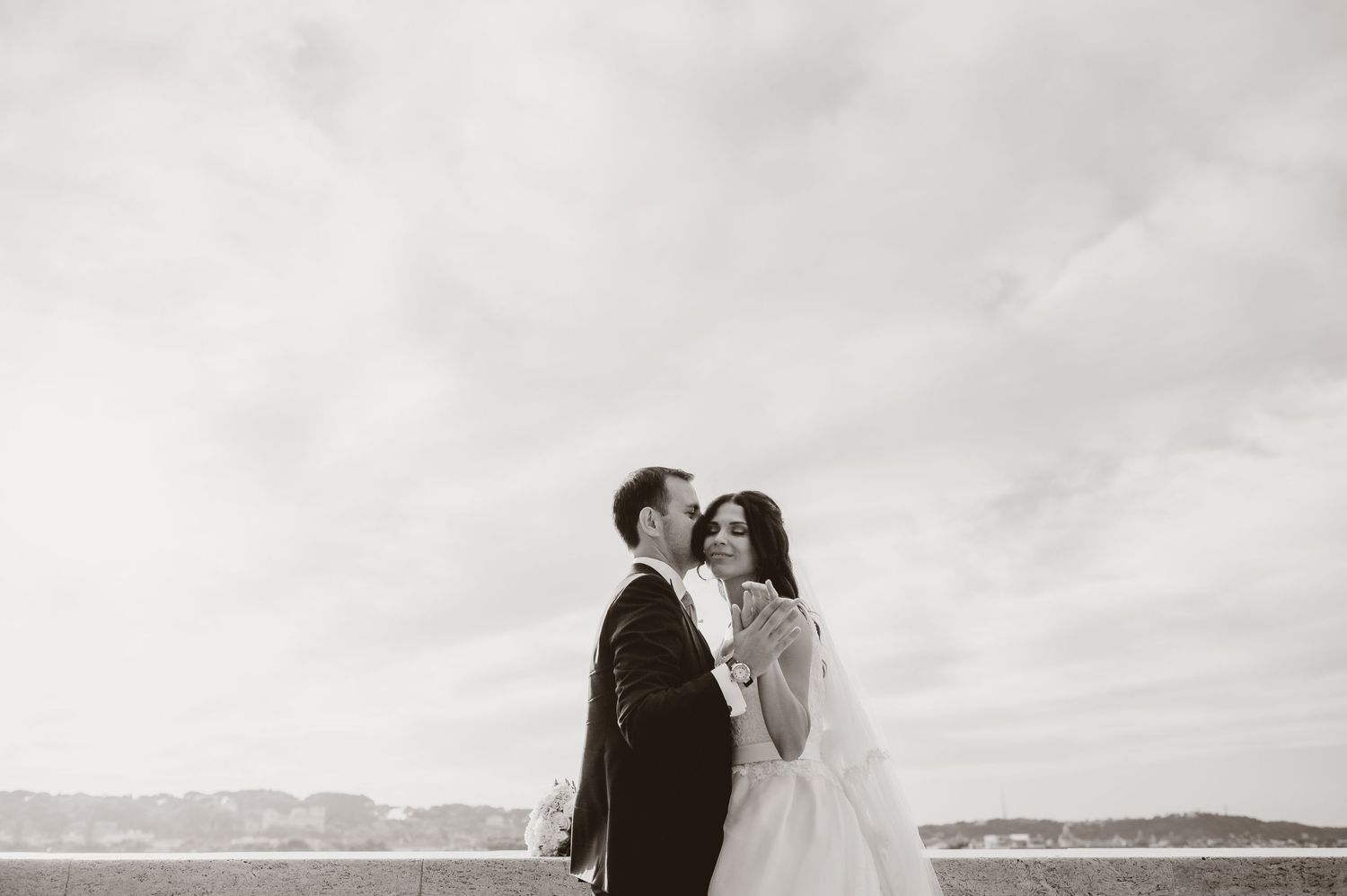 Wedding couple stands against dramatic cloudy sky overlooking mountain landscape.