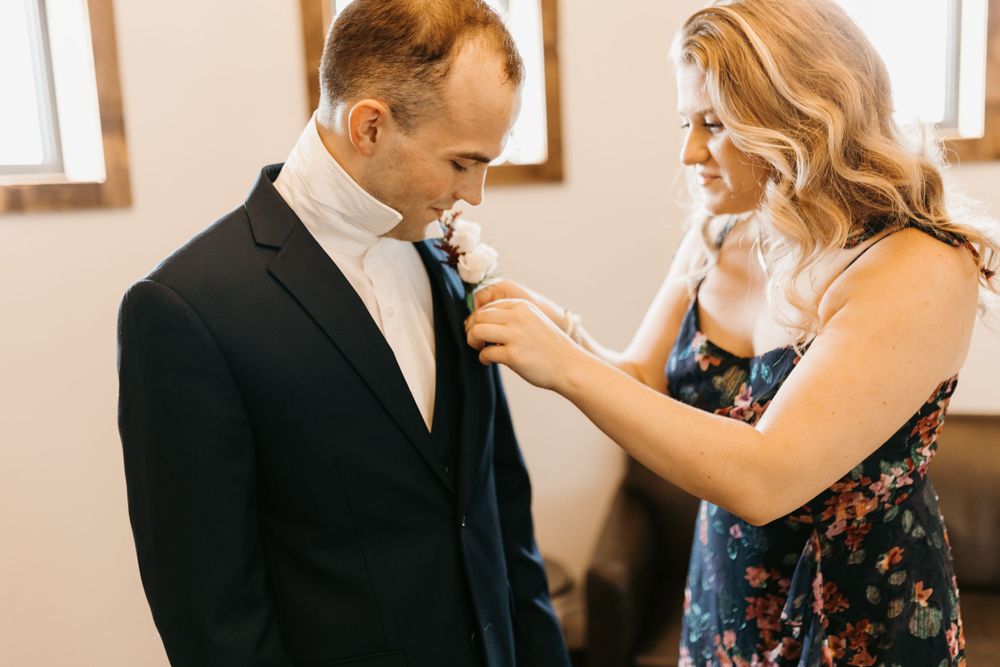 Woman in floral dress pinning white boutonniere on groom's navy suit jacket before wedding ceremony.