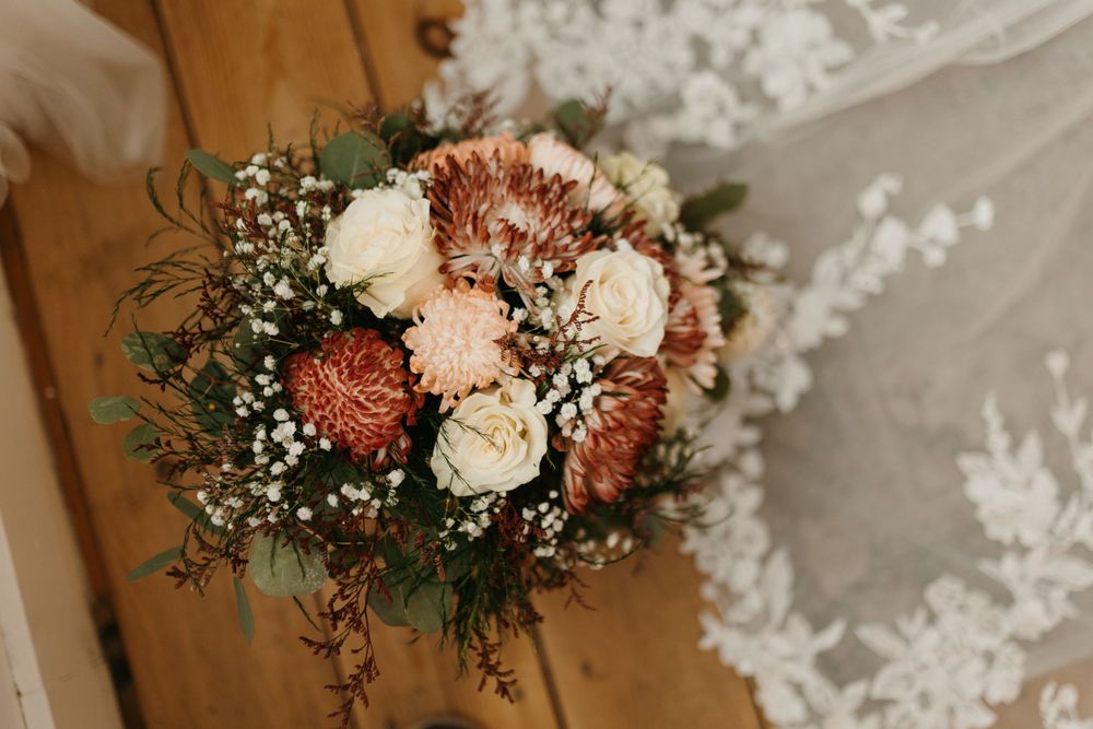 Rustic bridal bouquet with white roses, pink proteas, and baby's breath on wooden floor beside lace wedding dress.
