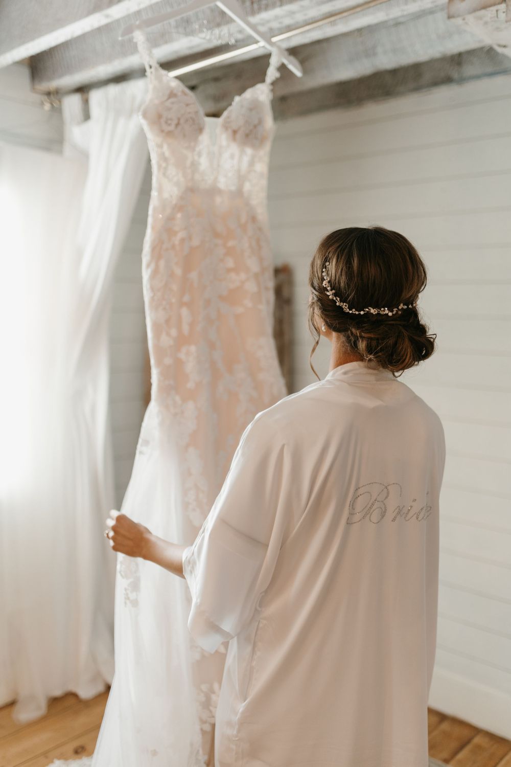 Bride in white robe with hair vine admiring lace wedding dress hanging in sunlit rustic room.