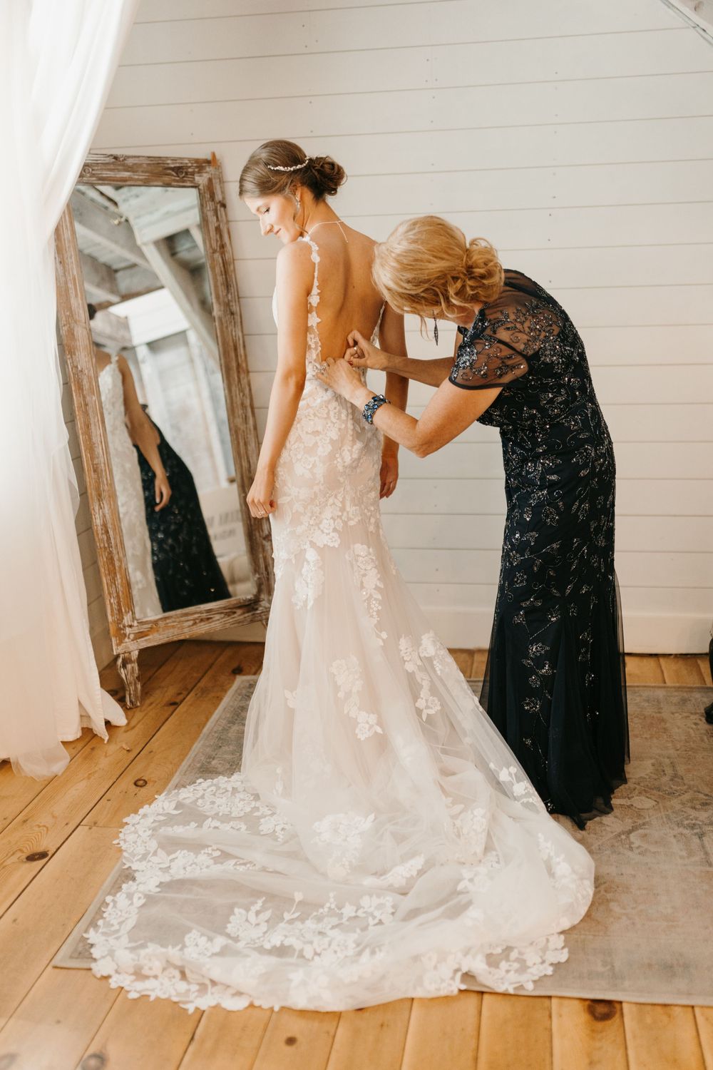 Woman in navy sequin dress fastening buttons on bride's lace backless wedding gown near vintage mirror.