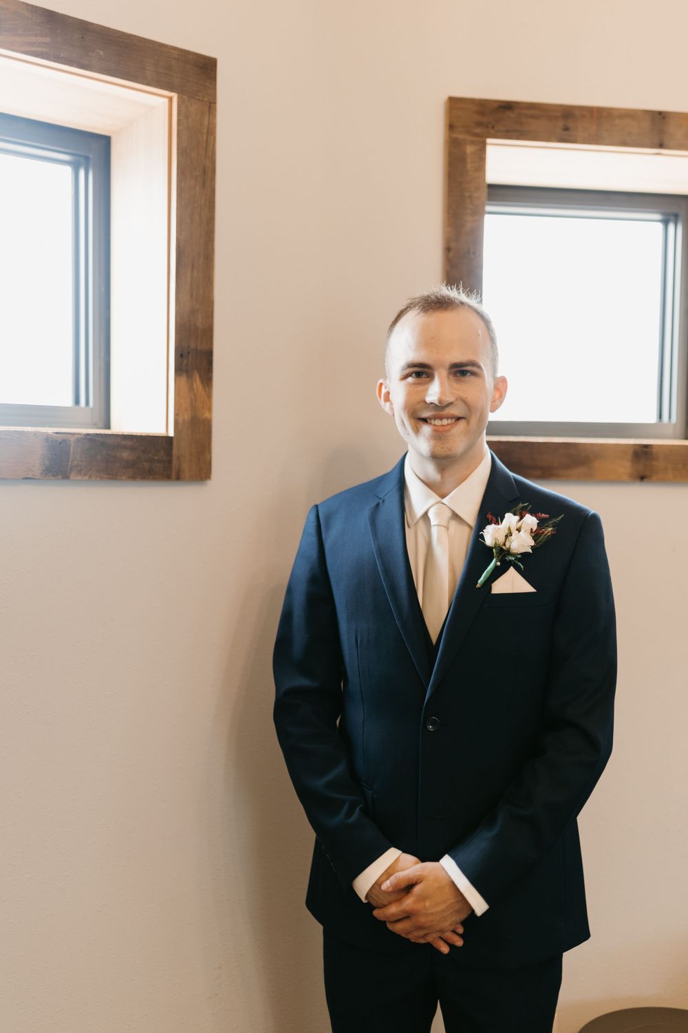 Smiling groom in navy suit with cream tie and floral boutonniere posing indoors.