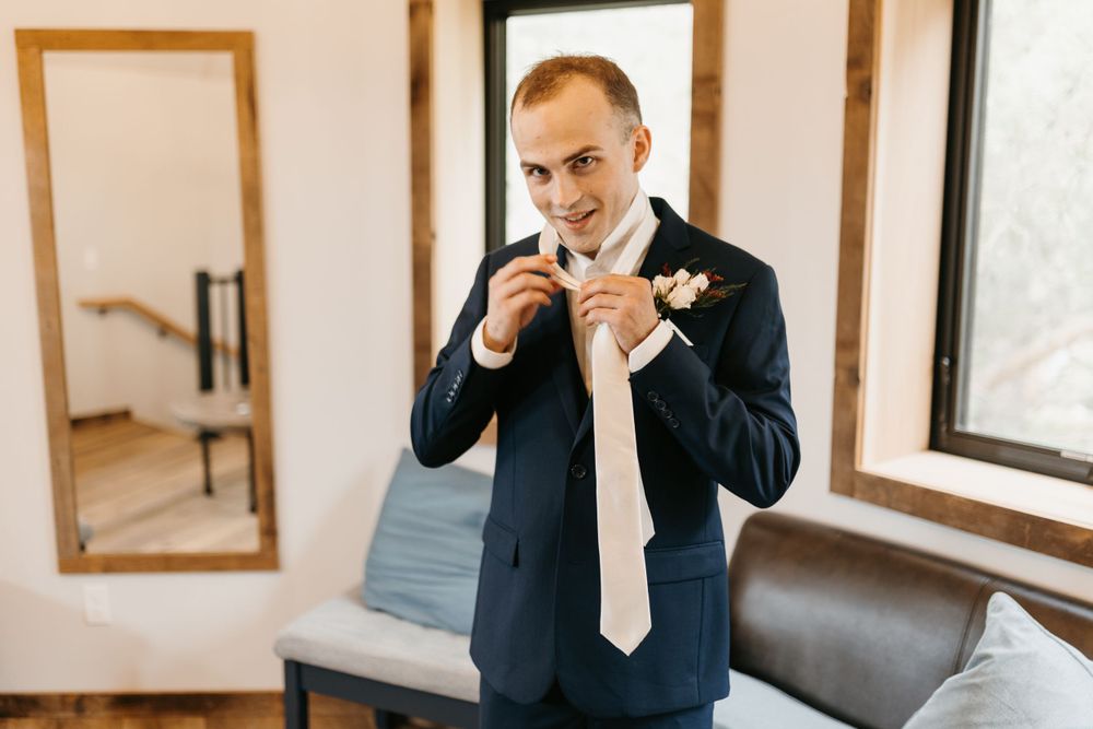 Groom smiling in navy suit tying cream necktie with floral boutonniere indoors.